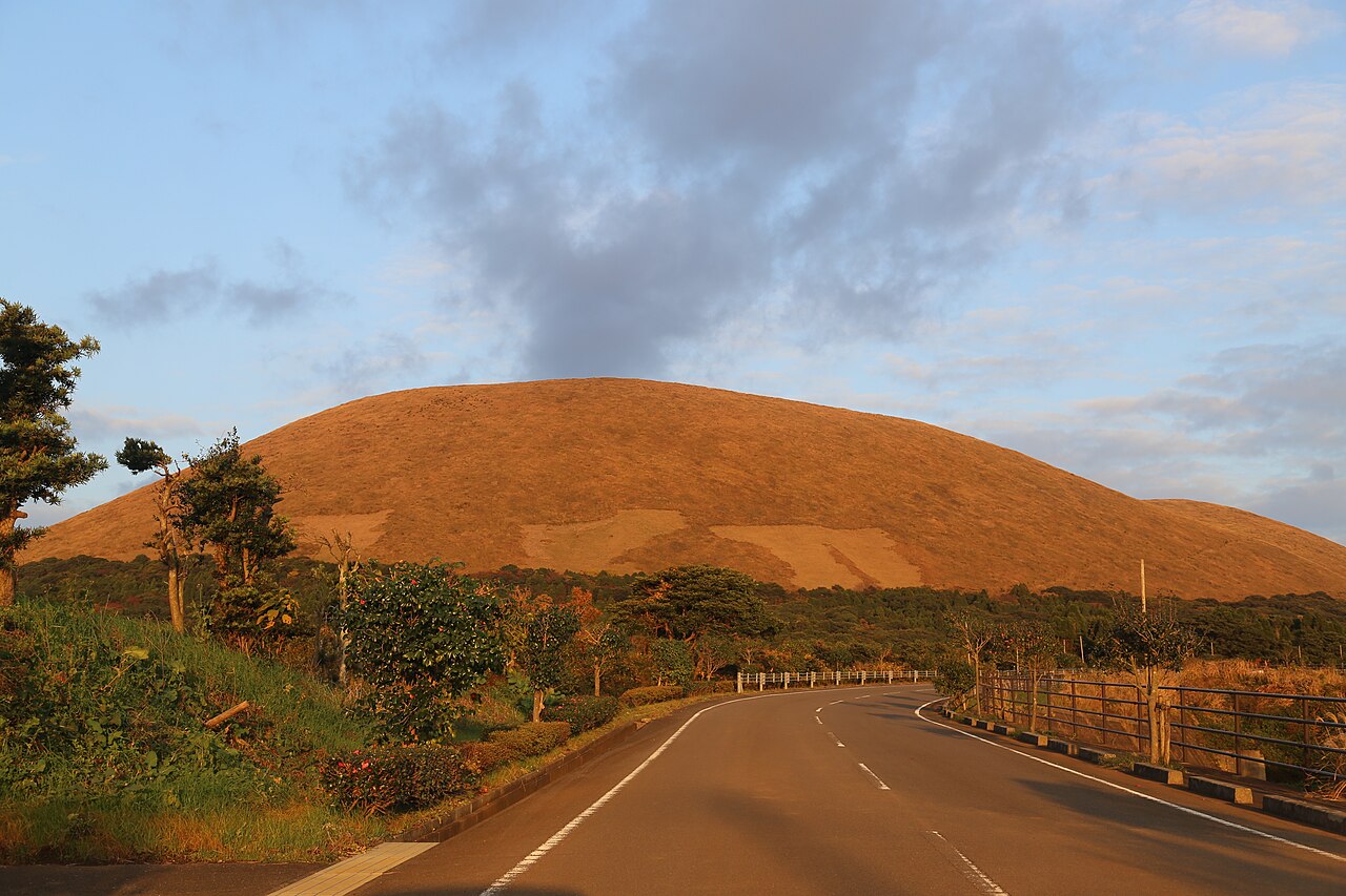 The grassy dome of Mount Onidake glowing gold at sunset, with an empty road curving through the foreground