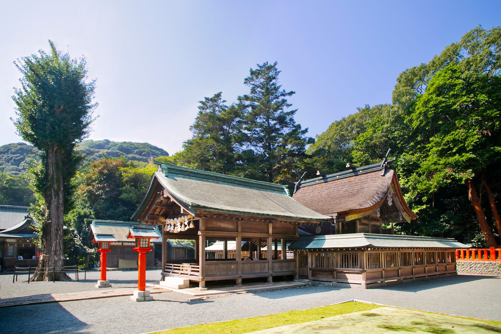 Shrine buildings of Nakatsumiya with bark-brown roofs and vermilion lanterns, set against green trees under a clear sky