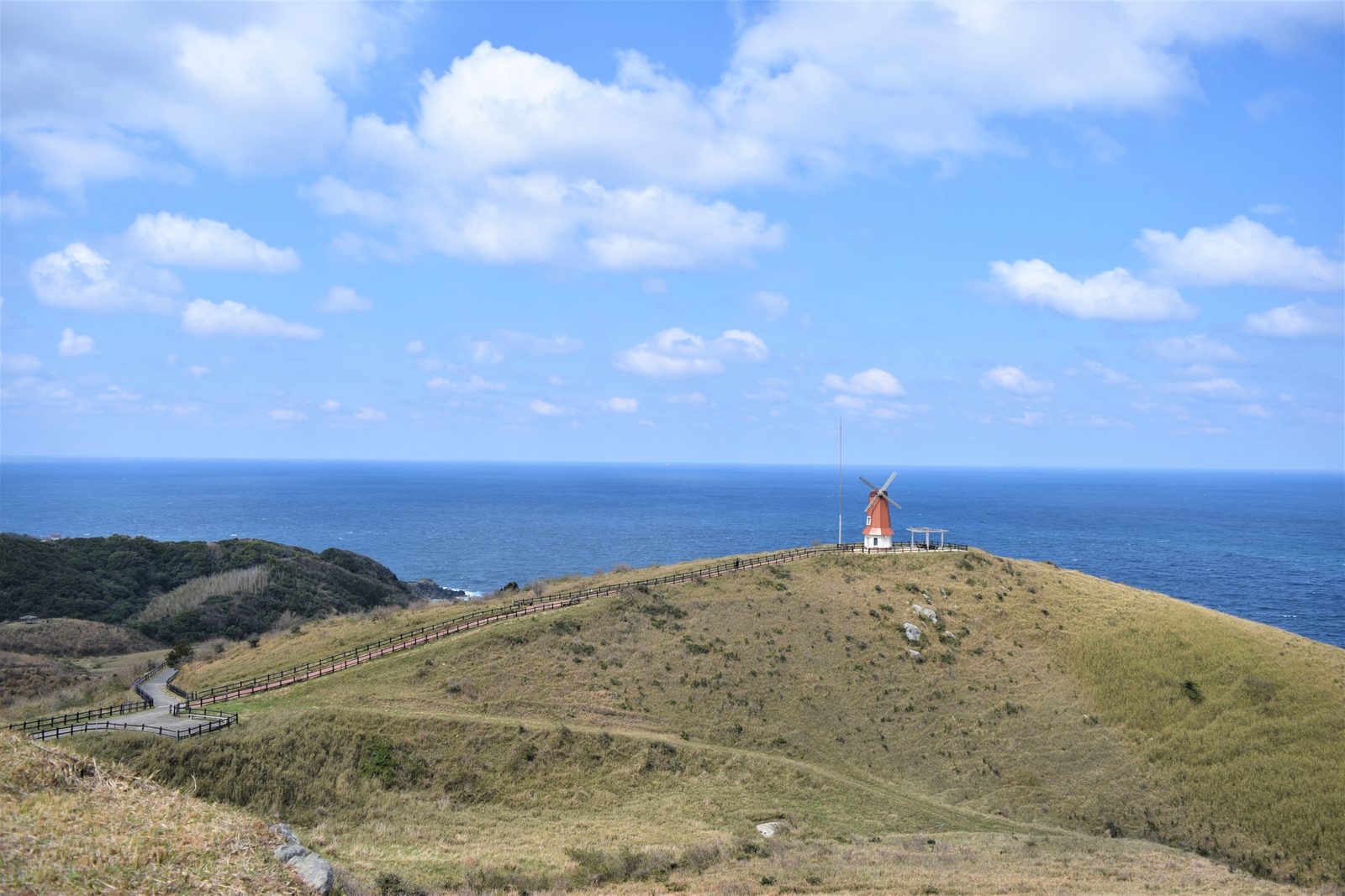 A red windmill on a grassy hill with the blue sea and sky behind it