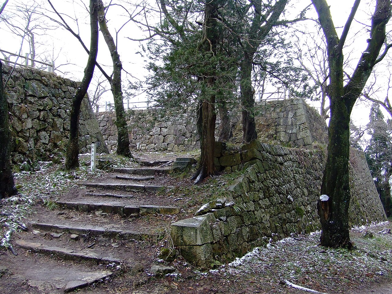 Worn stone steps climbing between moss-covered dry-stone ramparts on the wooded hill of Murakami Castle ruins, with bare winter trees and a dusting of snow on the ground.