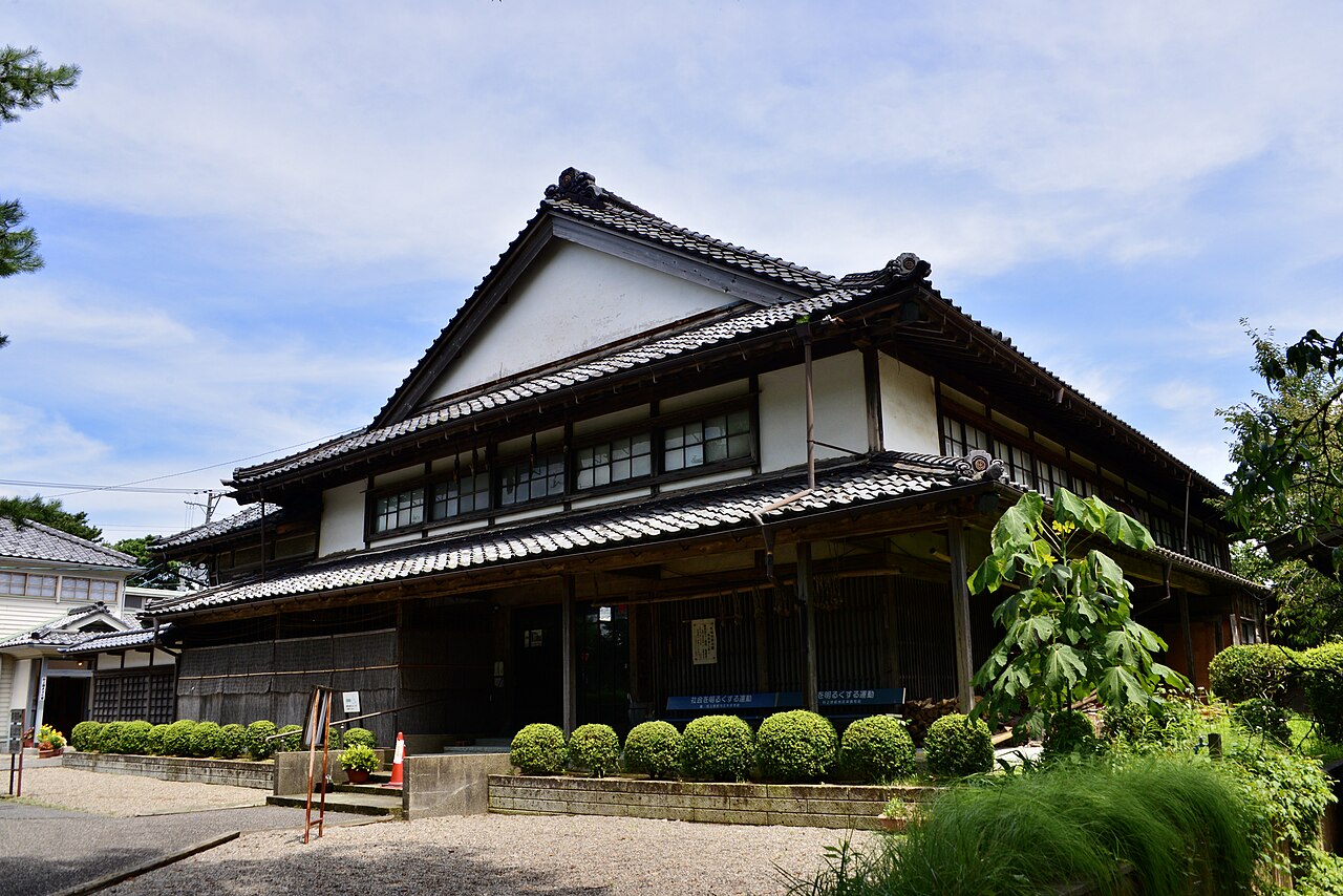A two-storey wooden merchant house in Murakami with white plaster gables, deep tiled eaves and a row of clipped shrubs along the gravel forecourt.
