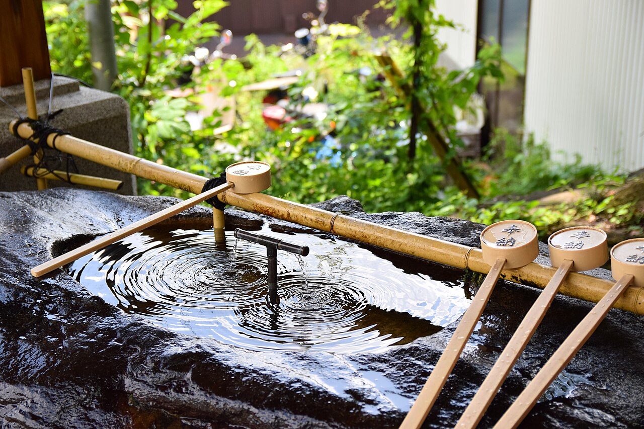 A stone water basin at Musashi Mitake Shrine, with bamboo ladles resting on a rail and ripples spreading across the still water.