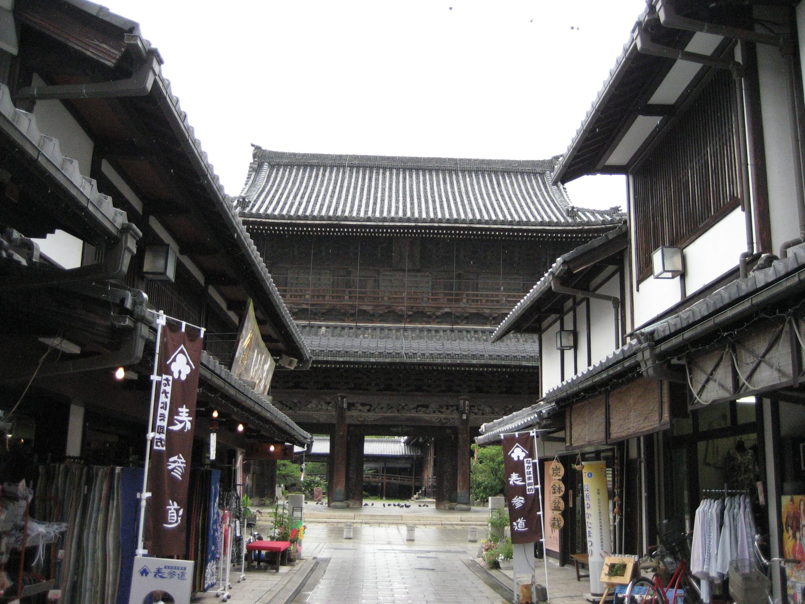 The gate of Daitsuji at the end of its approach street, with townhouses and banners on both sides under an overcast sky