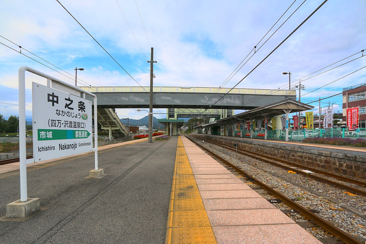 The single platform at Nakanojo under a bright sky, its name-board flanked by a covered footbridge and wooded hills.