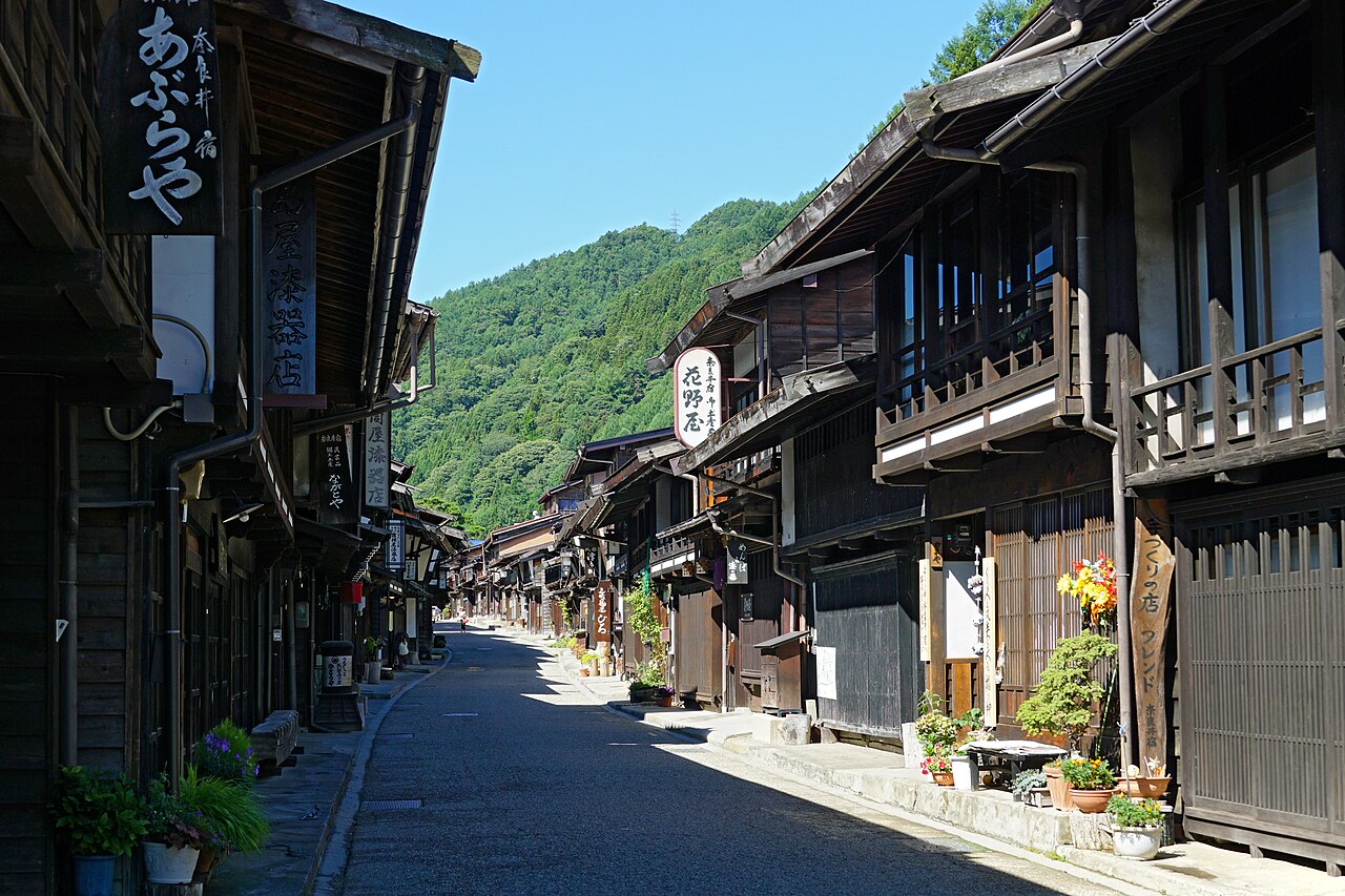 A street of dark timber townhouses with deep overhanging eaves and hanging shop signs at Narai-juku, running toward a forested ridge with no power poles in view.