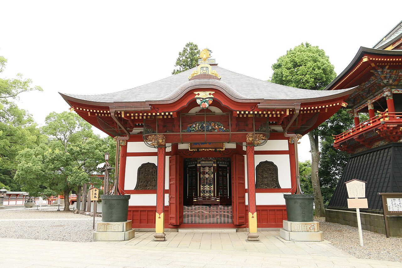 A hall of Naritasan Shinshoji Temple beside its three-storied pagoda, framed by trees.