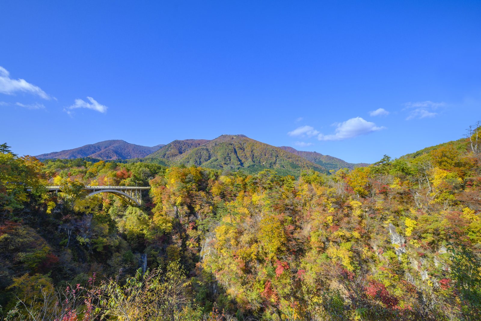 Ofukazawa Bridge arching over the autumn-colored cliffs of Naruko Gorge, with a forested mountain ridge and blue sky behind.
