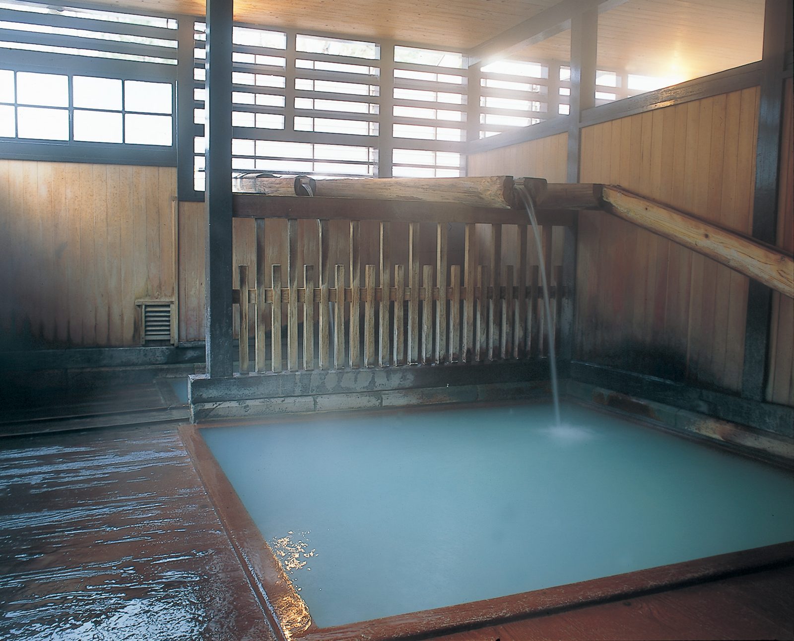 Empty indoor bathing pool at a Naruko Onsen public bathhouse, with pale blue-white hot spring water, a wooden slatted fence, and water flowing from a wooden spout.