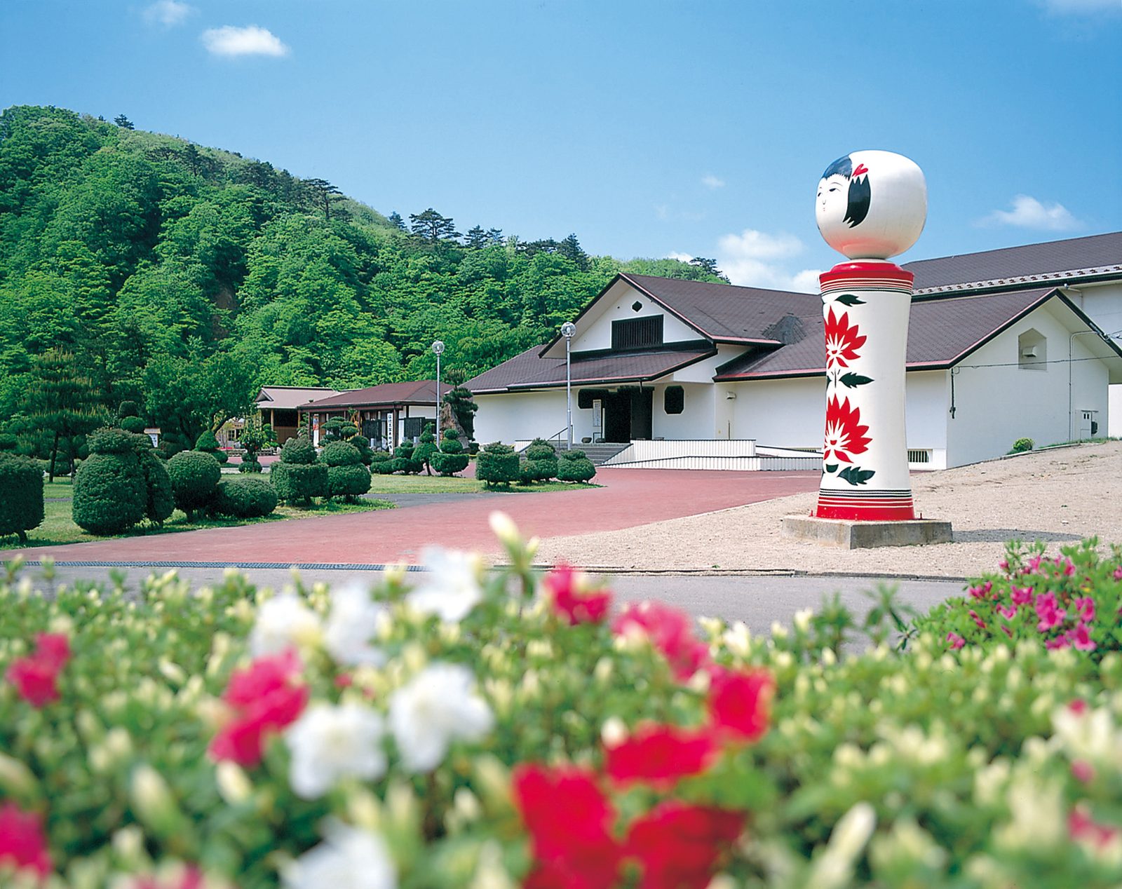 Giant kokeshi doll statue with a painted chrysanthemum pattern standing in front of the Japan Kokeshi Museum building in Naruko, with red and white azaleas blooming in the foreground.
