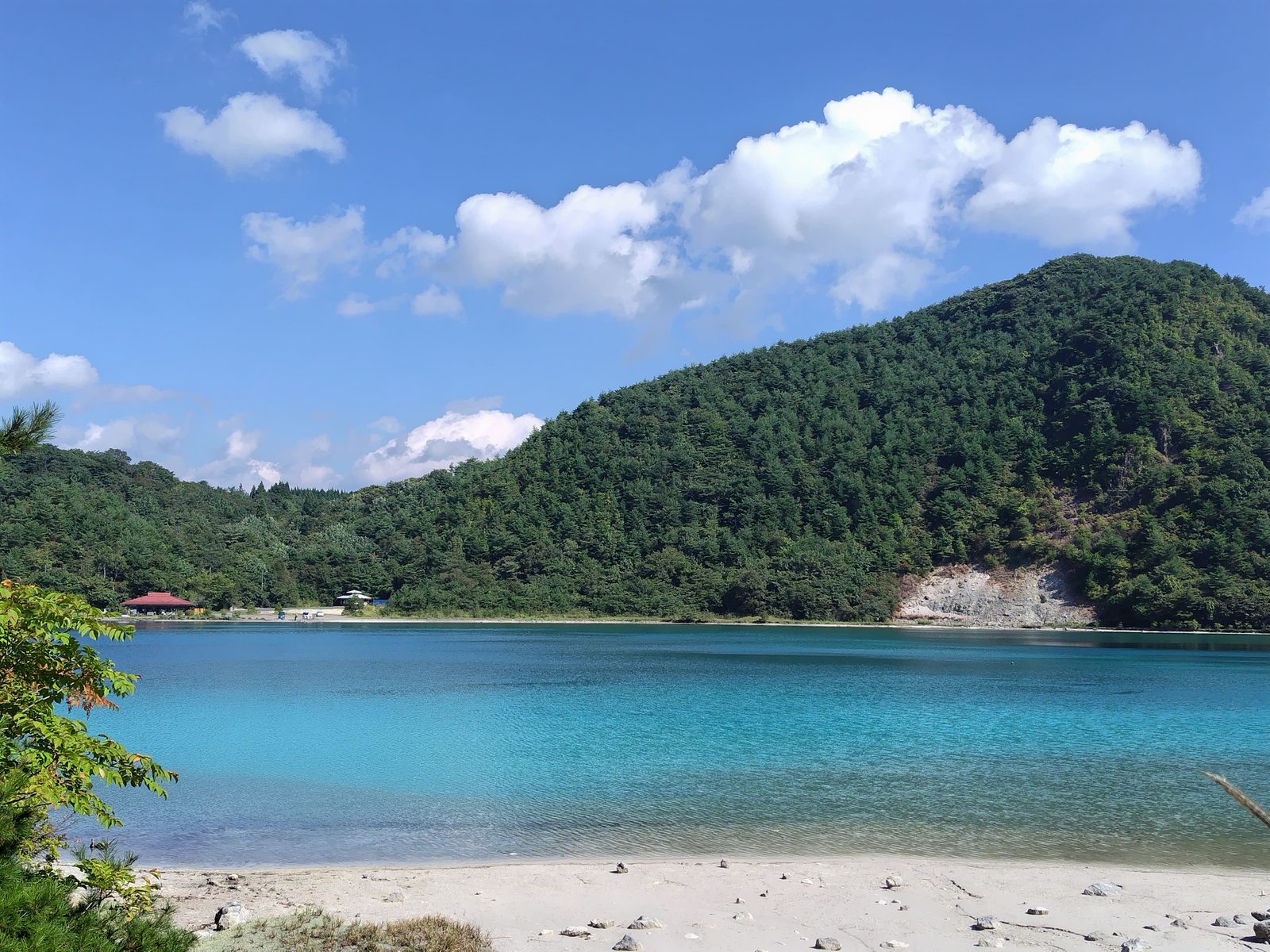 Turquoise crater lake at Katanuma near Naruko, with a pale sandy shore, pine-covered hillside, and blue sky with clouds.