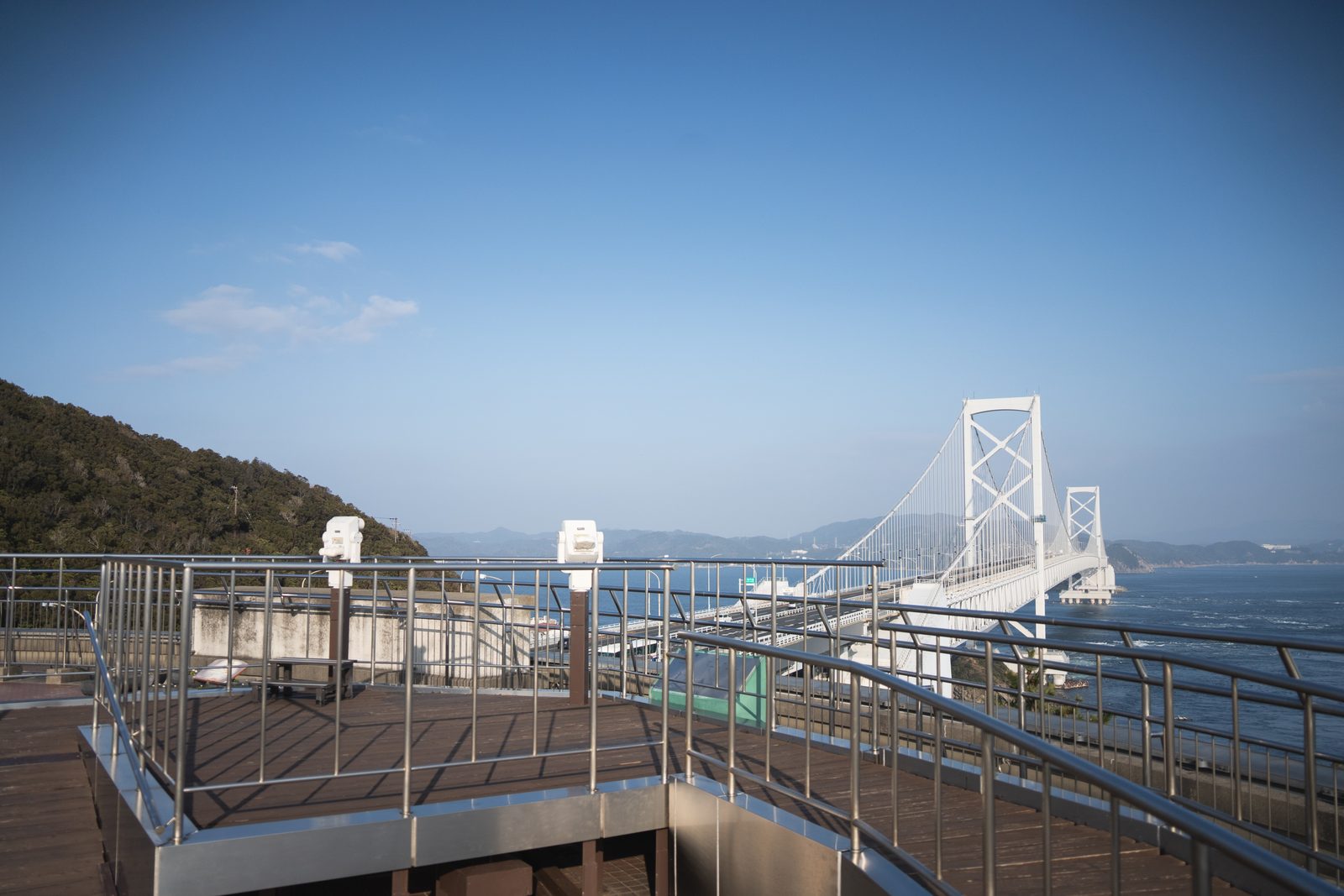 A wooden observation deck with steel railings and coin-operated binoculars looking toward the white Onaruto Bridge across blue water.