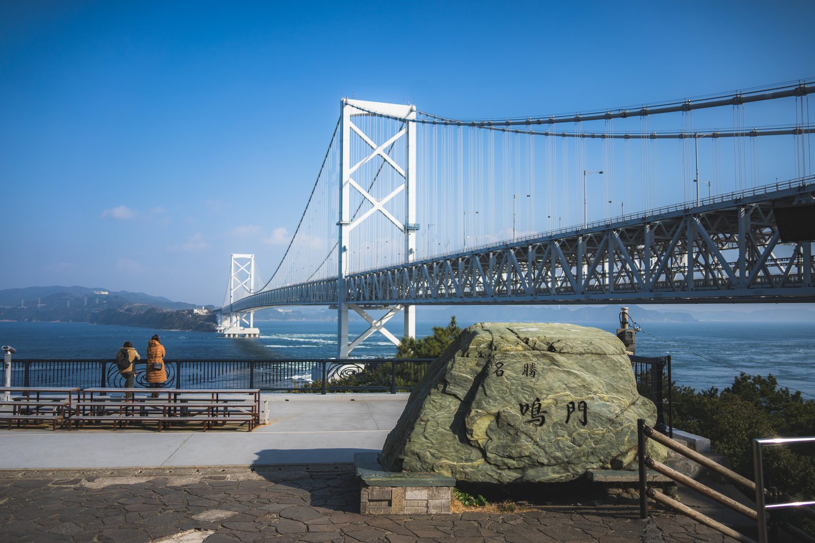 A carved stone monument reading Naruto stands on a paved lookout, with the long steel truss of the Onaruto Bridge stretching across the strait behind it and two visitors at the railing.