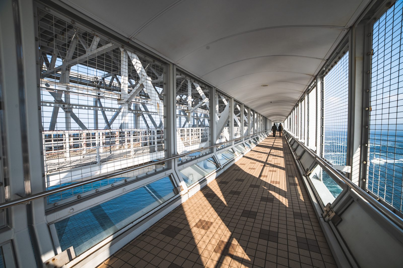 A long enclosed walkway with a tiled floor and mesh windows leading out over the Naruto Strait, with two visitors far ahead.