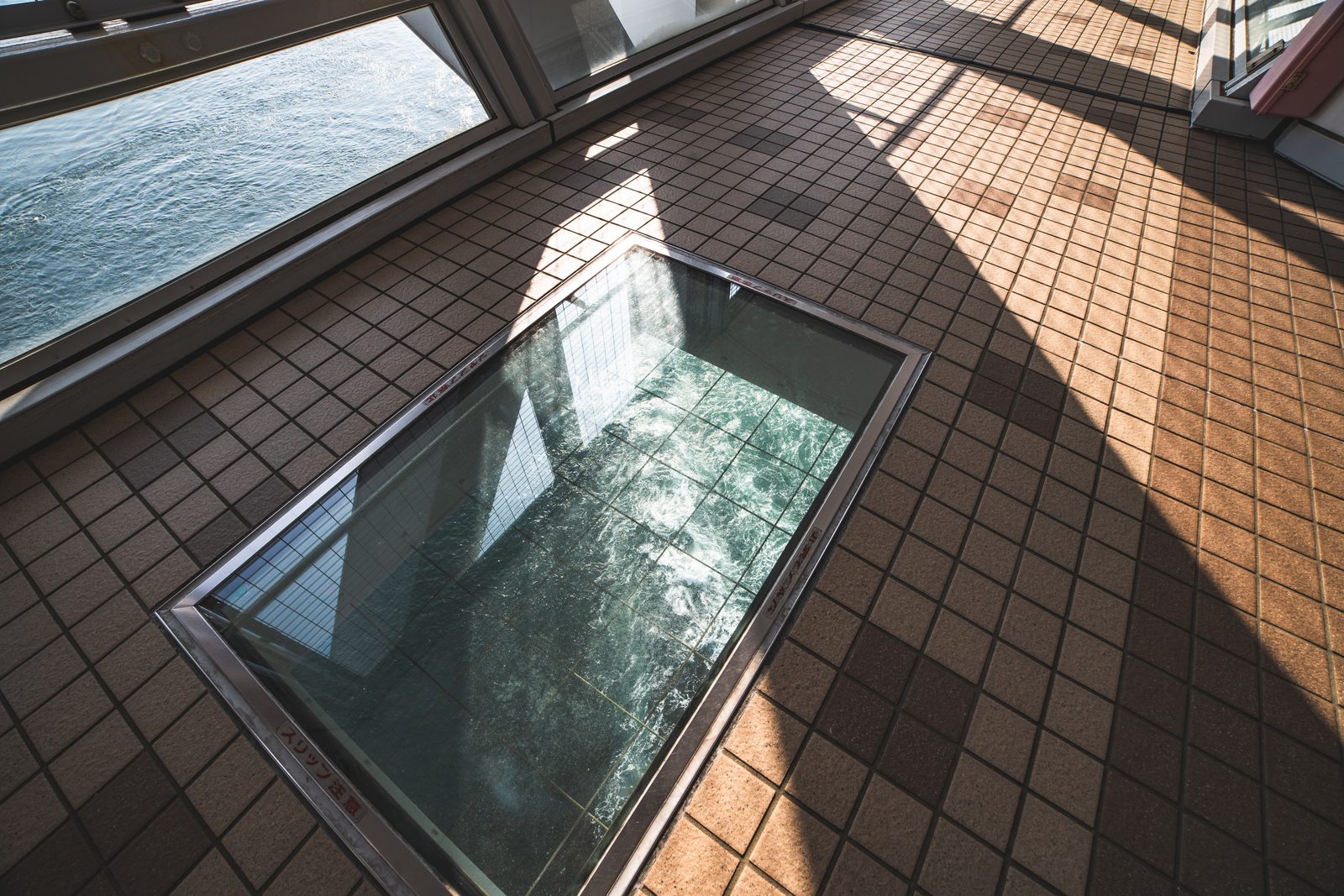 A rectangular glass panel set into a tiled walkway floor, showing churning green water far below.