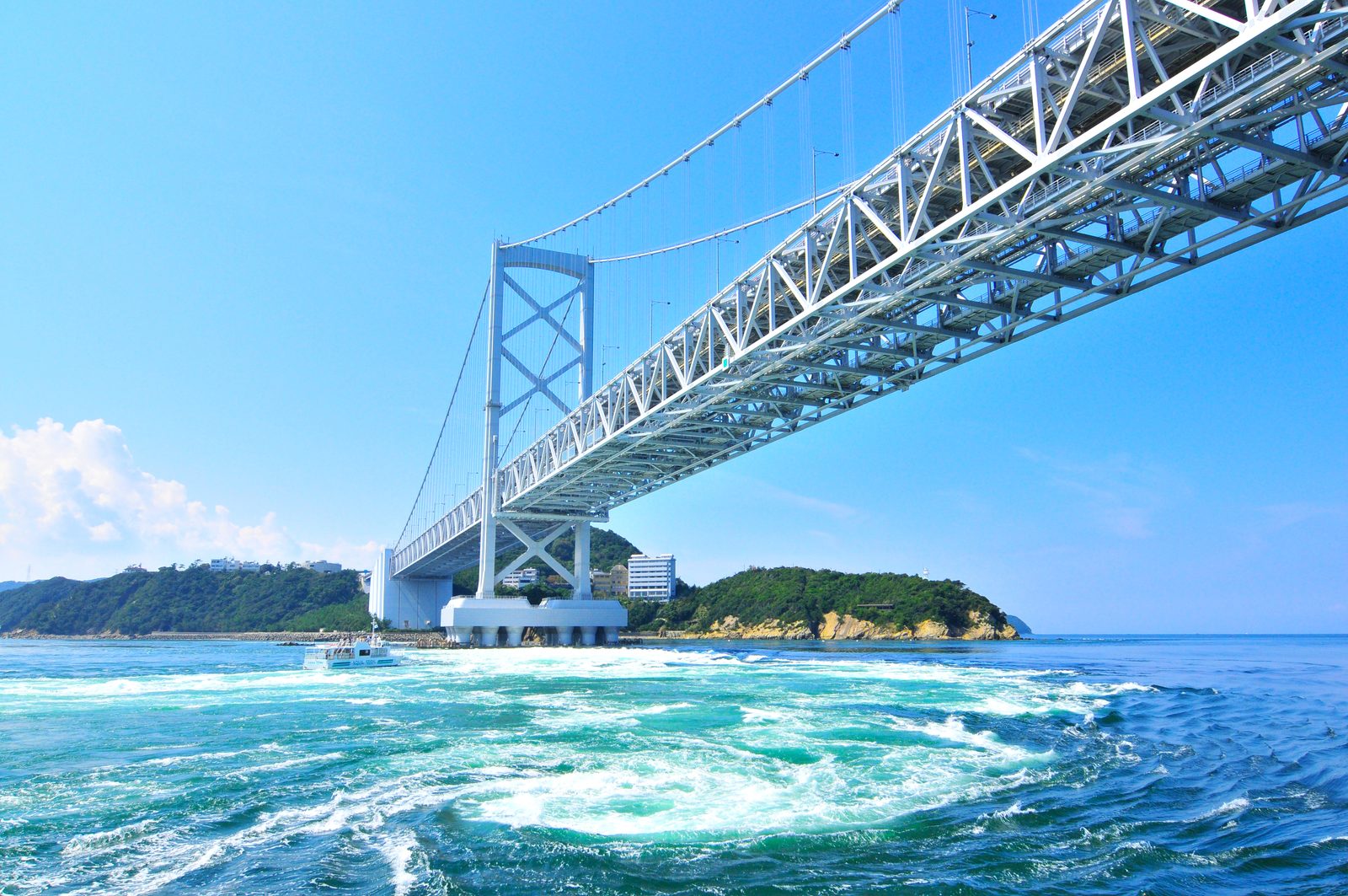 The Onaruto Bridge seen from the water, with churning white and green currents in the foreground and a small boat near the far tower.