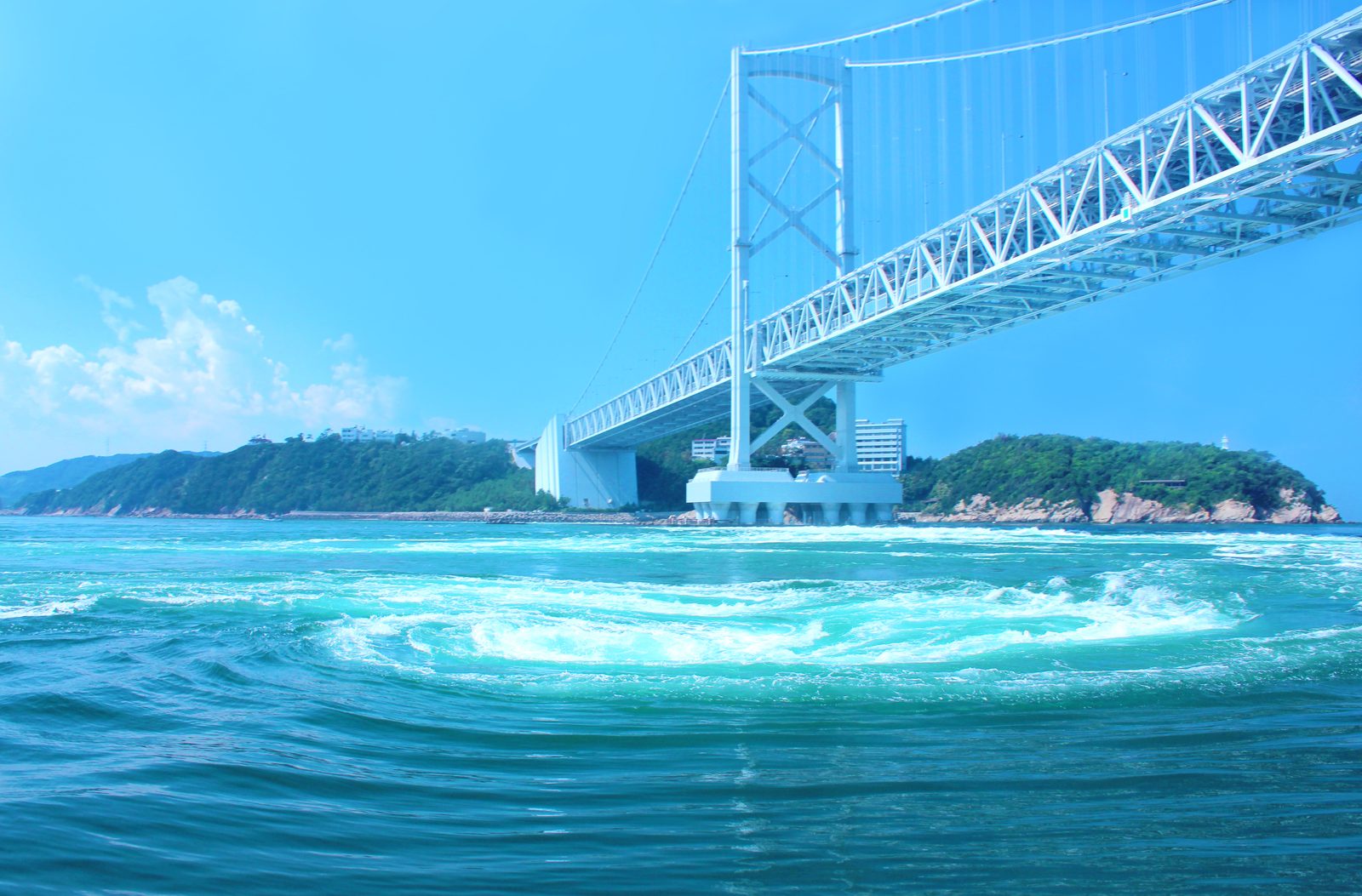 Pale turquoise whirlpools swirling in the strait below the white towers and steel deck of the Onaruto Bridge, with a green rocky island beyond.