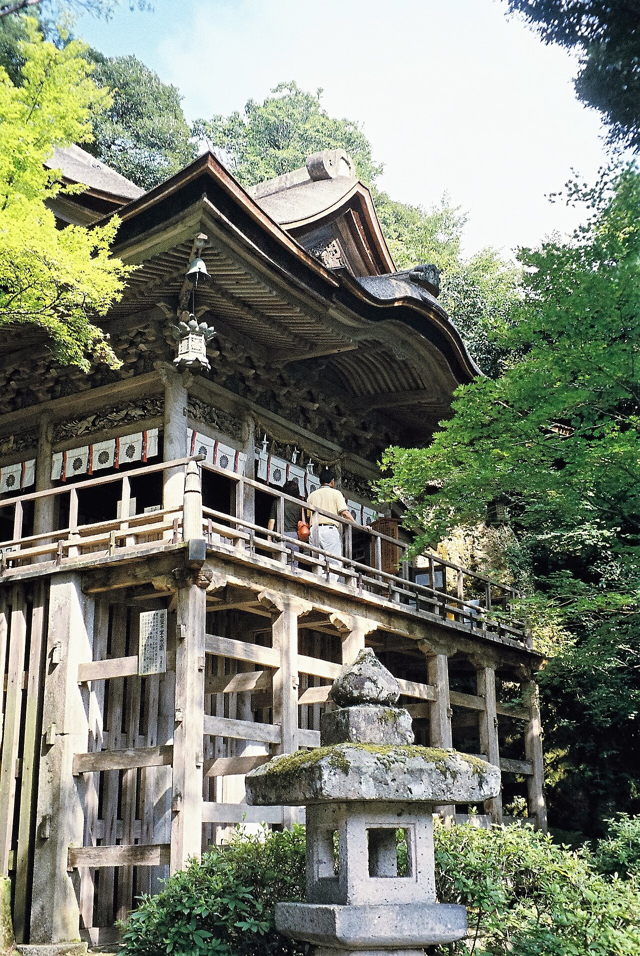 A timber temple hall raised on tall wooden pillars stands against a rugged rock face at Natadera, with a mossy stone lantern in the foreground and visitors on the walkway.