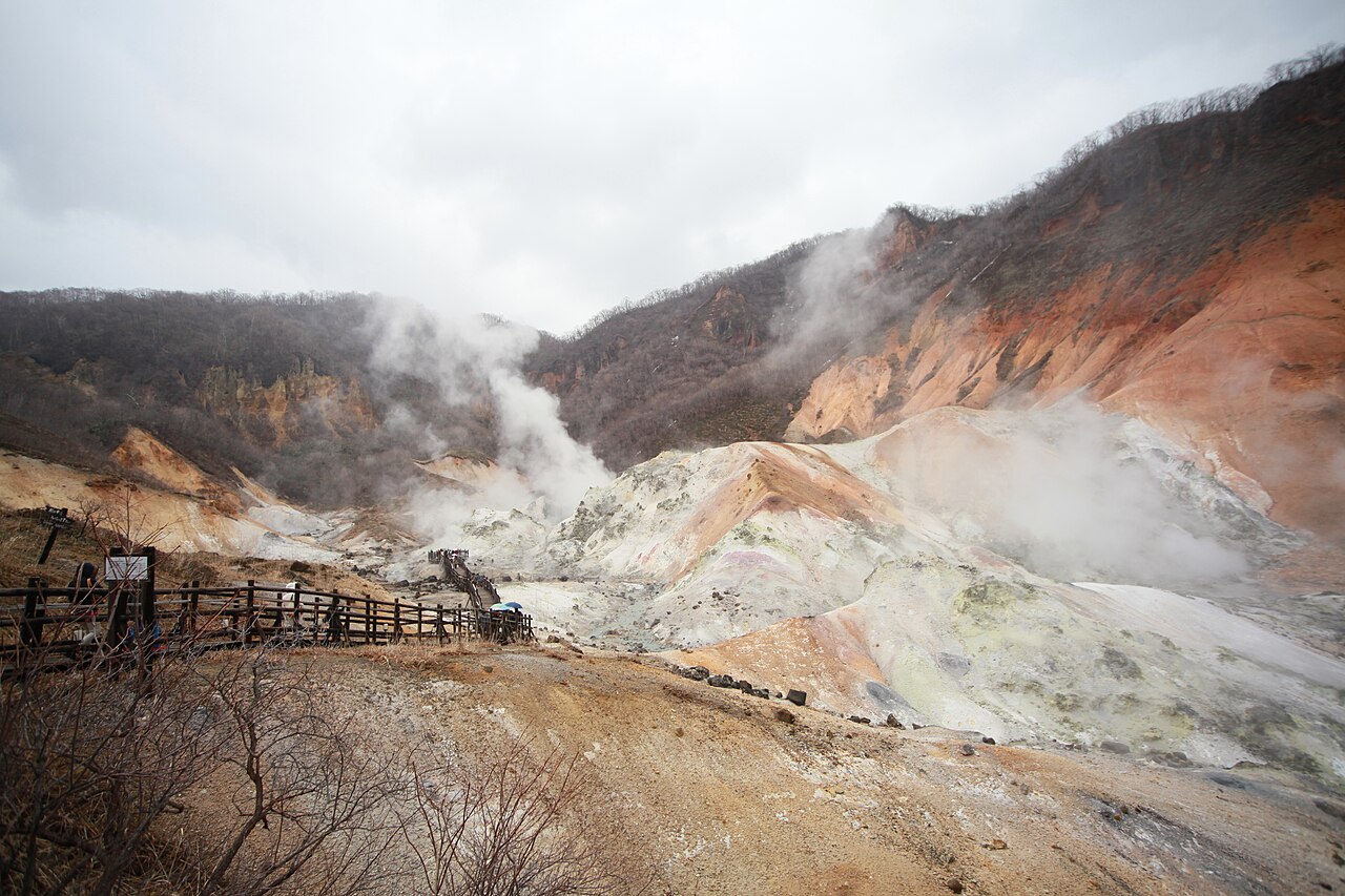 Steam rising from the ochre and pale-grey slopes of Noboribetsu Jigokudani, with a wooden railed boardwalk running along the left edge of the crater floor.