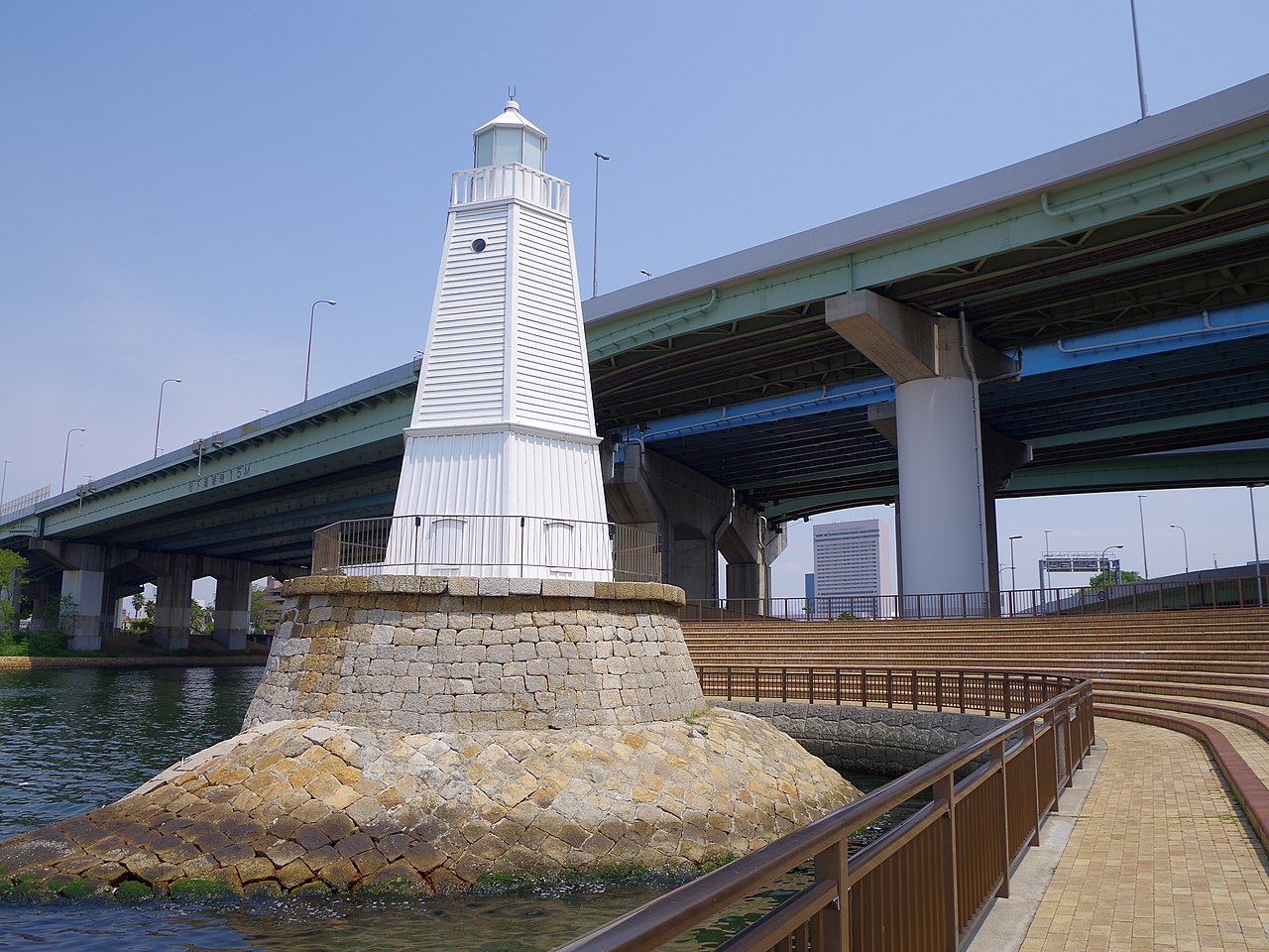 The white wooden hexagonal form of the old Sakai lighthouse on its stone base at the water's edge, with an elevated expressway behind