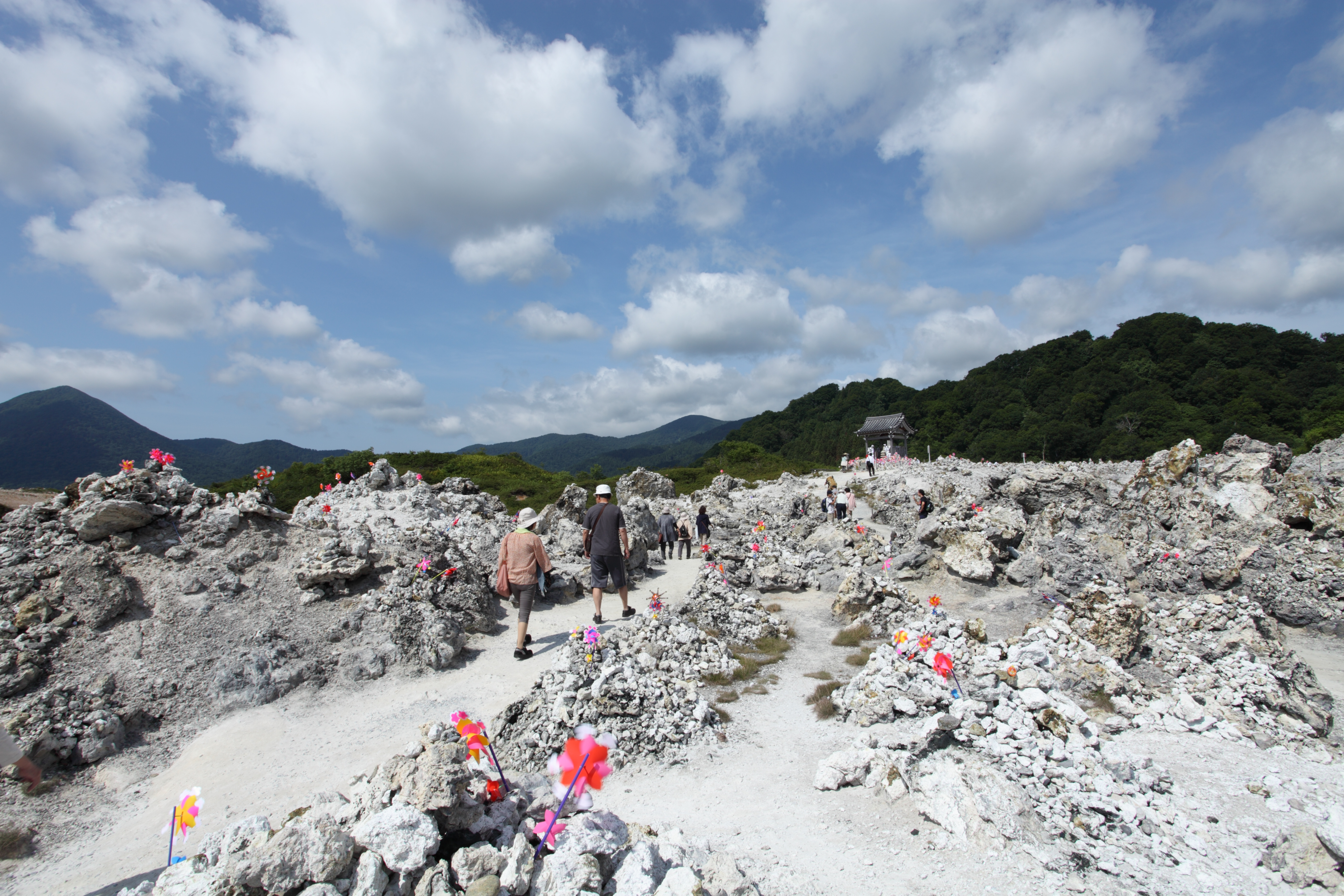Visitors walking a path through pale, rocky volcanic ground with piles of stones and colorful pinwheels under a blue sky with clouds