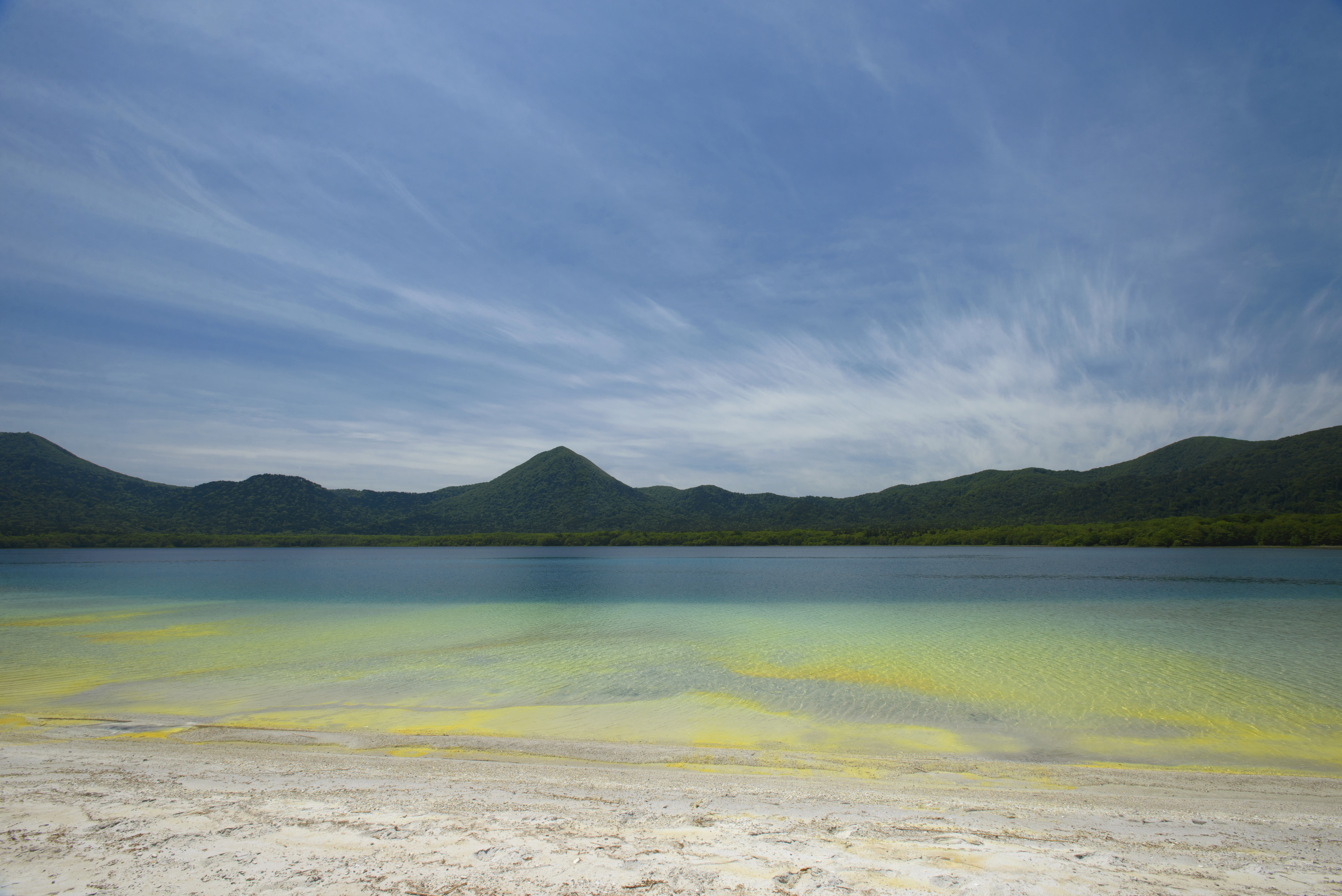 A calm lake with turquoise and yellow-green shallows, a sandy shore and a forested hill beyond under streaked clouds