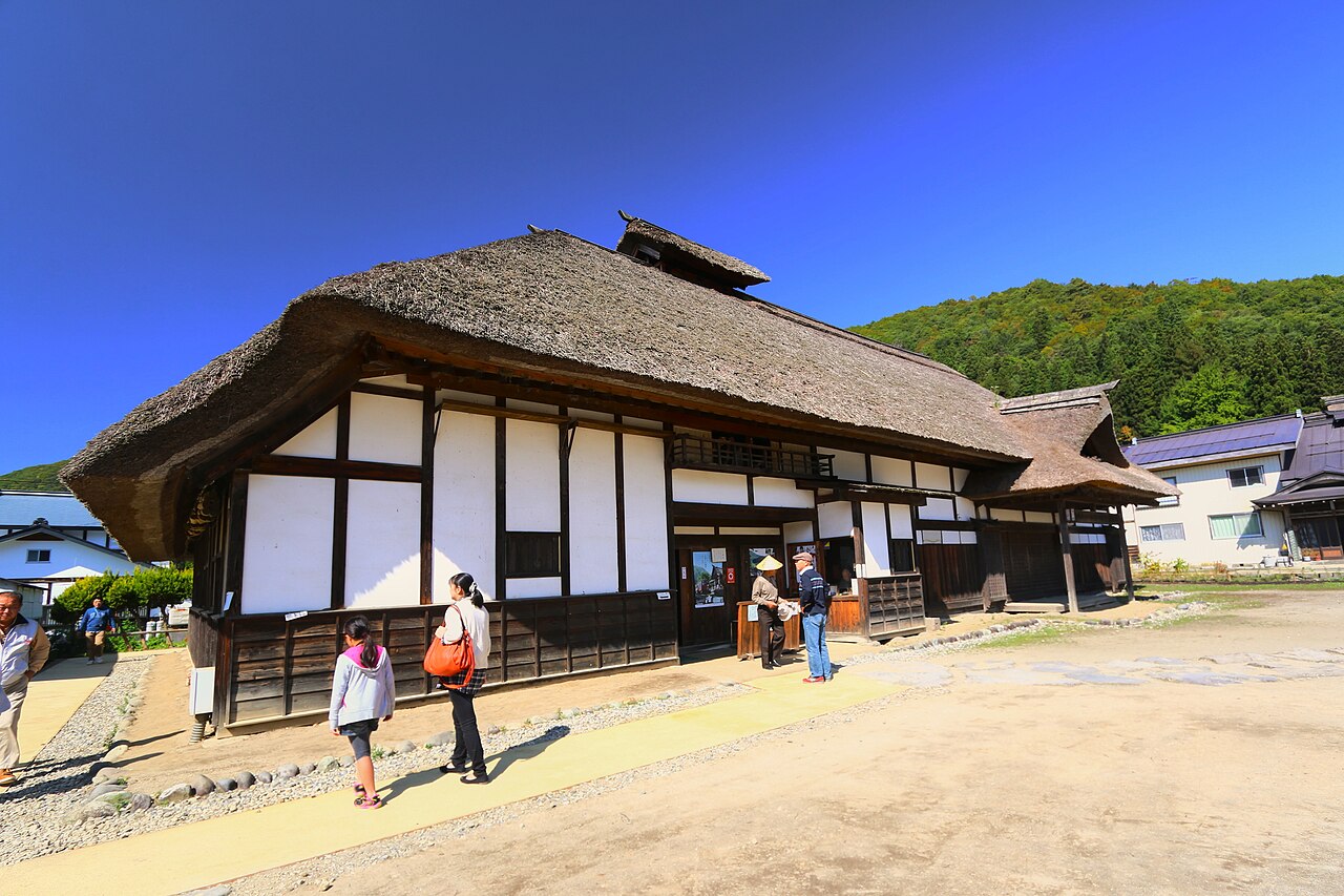 A large thatched-roof house with white walls stands beside the old post road in Ouchi-juku.