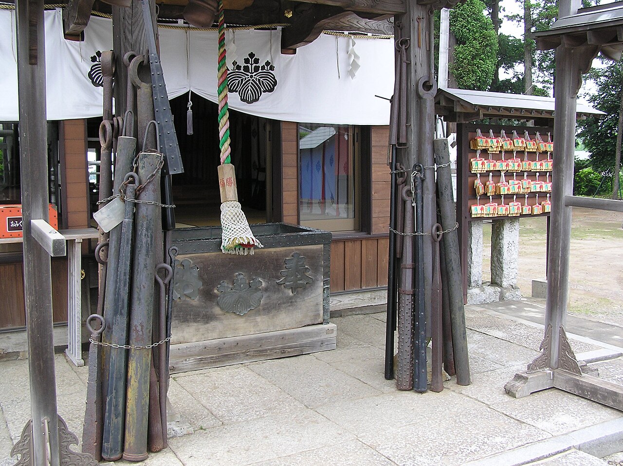 Heavy iron clubs stand chained against the pillars of the votive hall at Kijin Shrine in Ranzan, beside a white curtain and a rack of wooden prayer plaques.