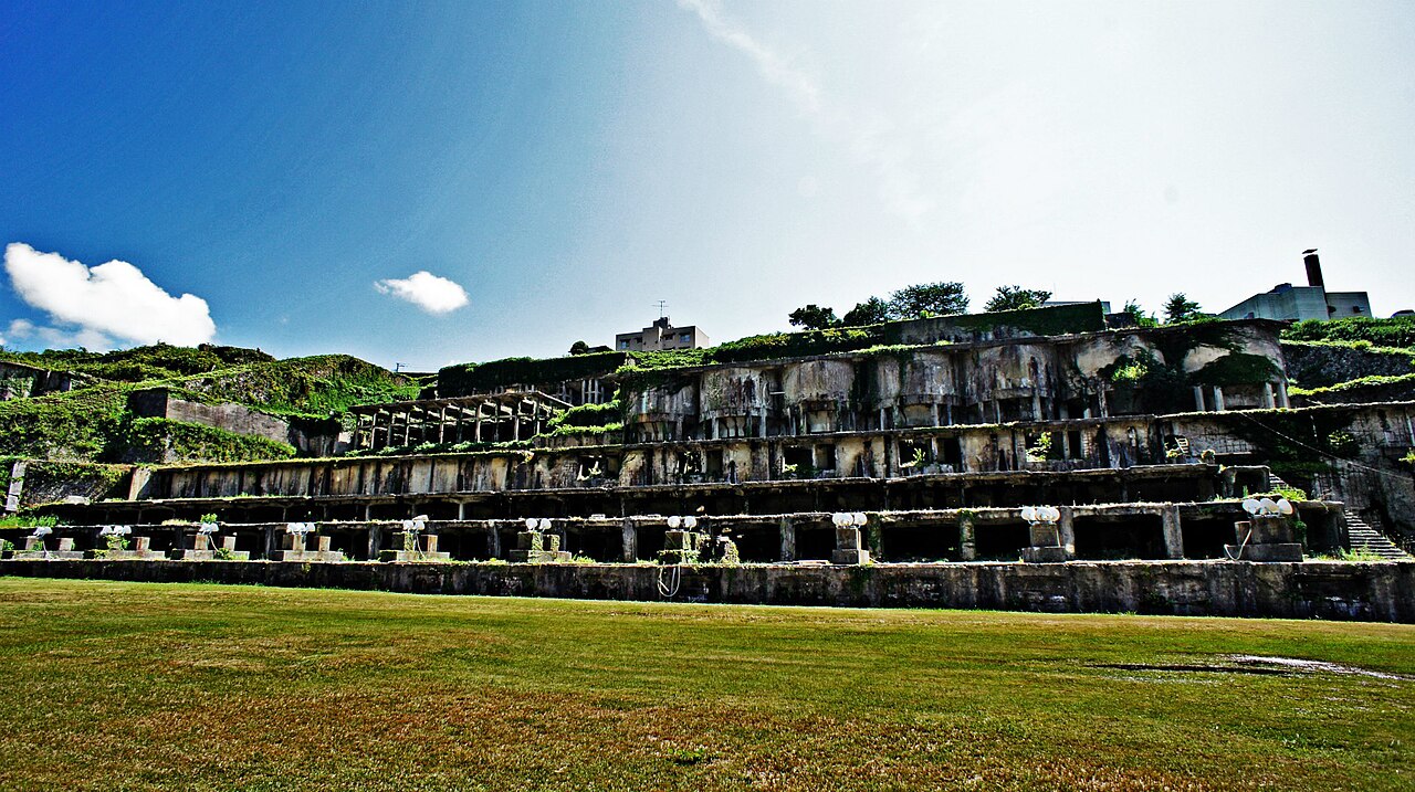Terraced concrete ruins of a former ore-processing plant, overgrown with grass, rise above an open lawn under a blue sky at the Sado mine site.