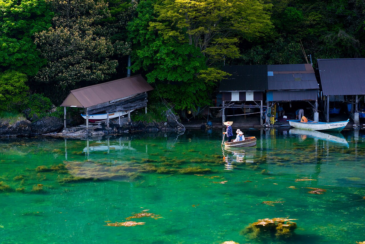 Two people in a round wooden tub boat cross clear green water past weathered wooden boat sheds below a forested slope on the Sado coast.