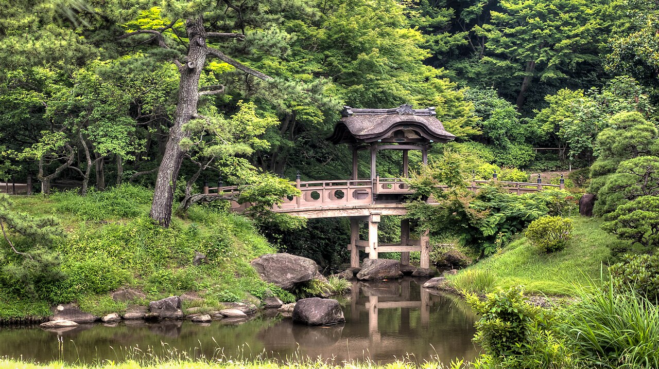 A thatched-roof covered bridge crosses a rock-lined pond among pine and maple trees at Sankeien Garden, its reflection visible in the still water.
