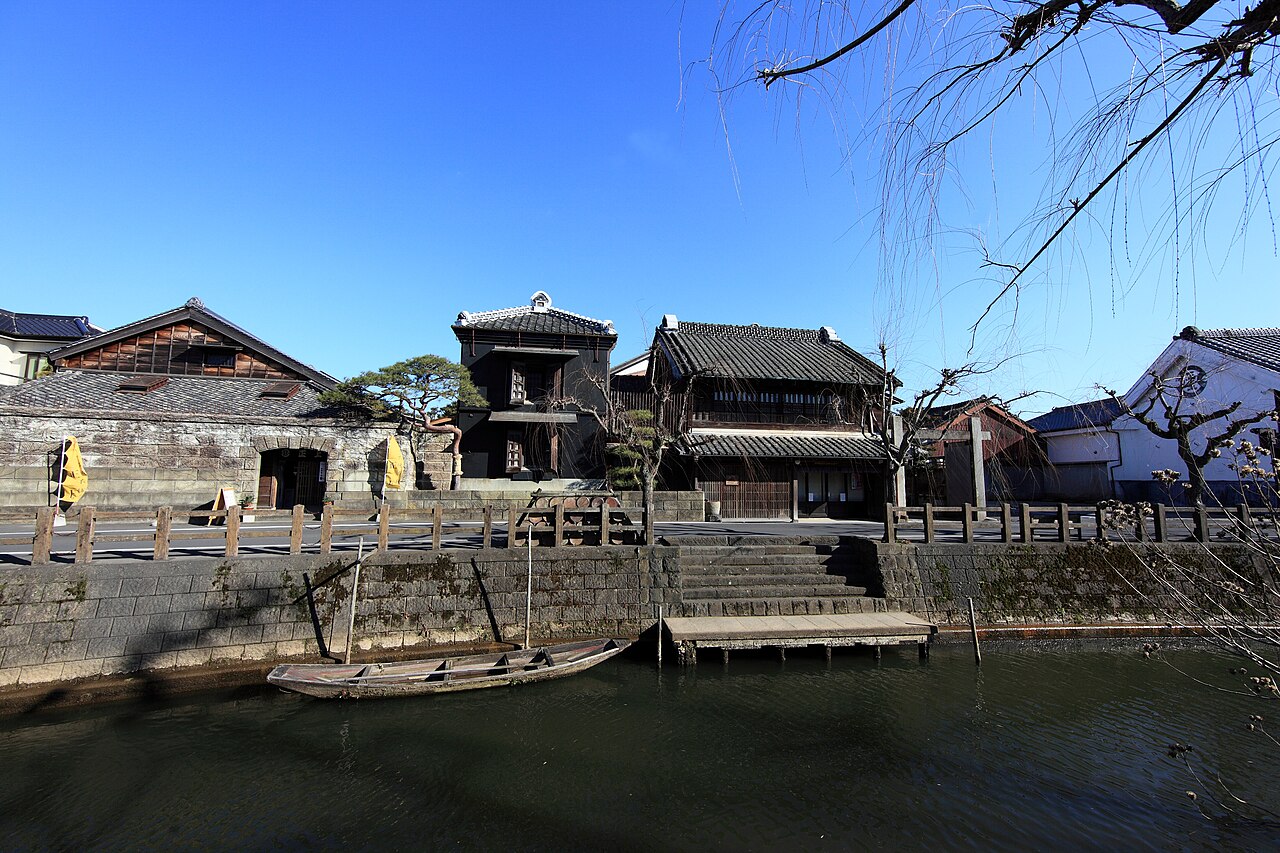 Black-walled Edo-period merchant houses and a moored wooden boat along the stone-lined Ono River in Sawara, under a clear blue sky.
