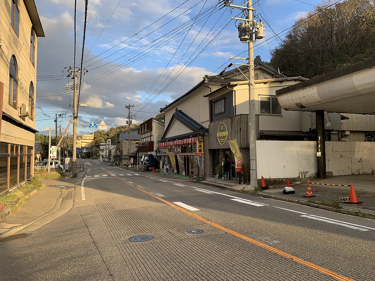 A quiet street through Senami Onsen in late afternoon light, lined with a tiled shopfront under a striped awning, hanging banners and overhead wires, with wooded slopes rising behind.