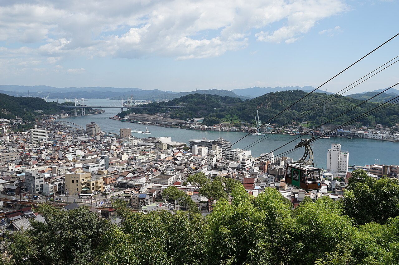 A ropeway car climbing above the rooftops of Onomichi, with the channel, the bridge and the island opposite spread out below.