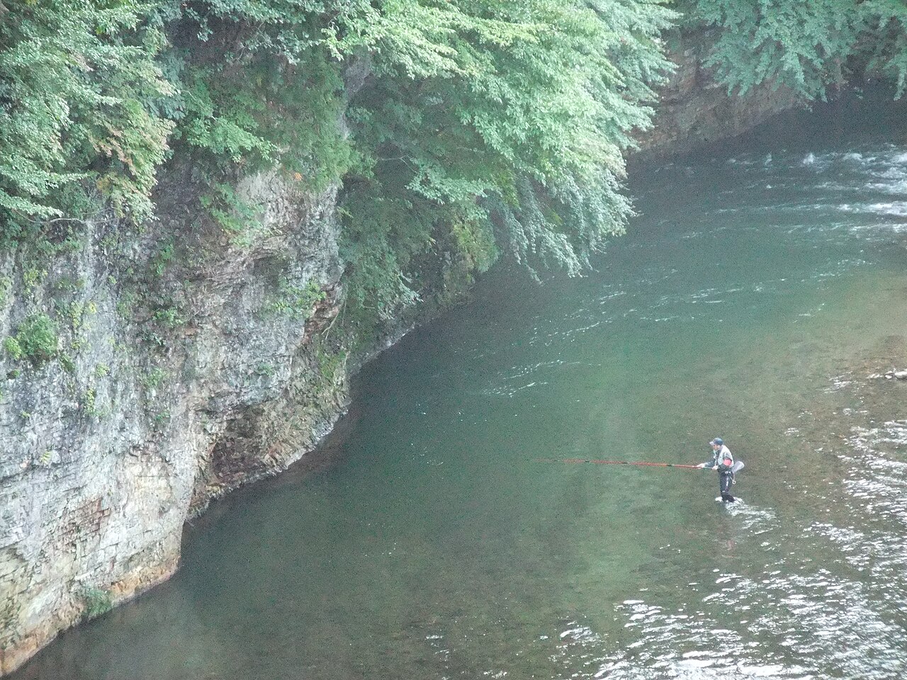 A lone angler standing in the clear green water of the Toyosawa River below a mossy rock face in the Shidotaira gorge.