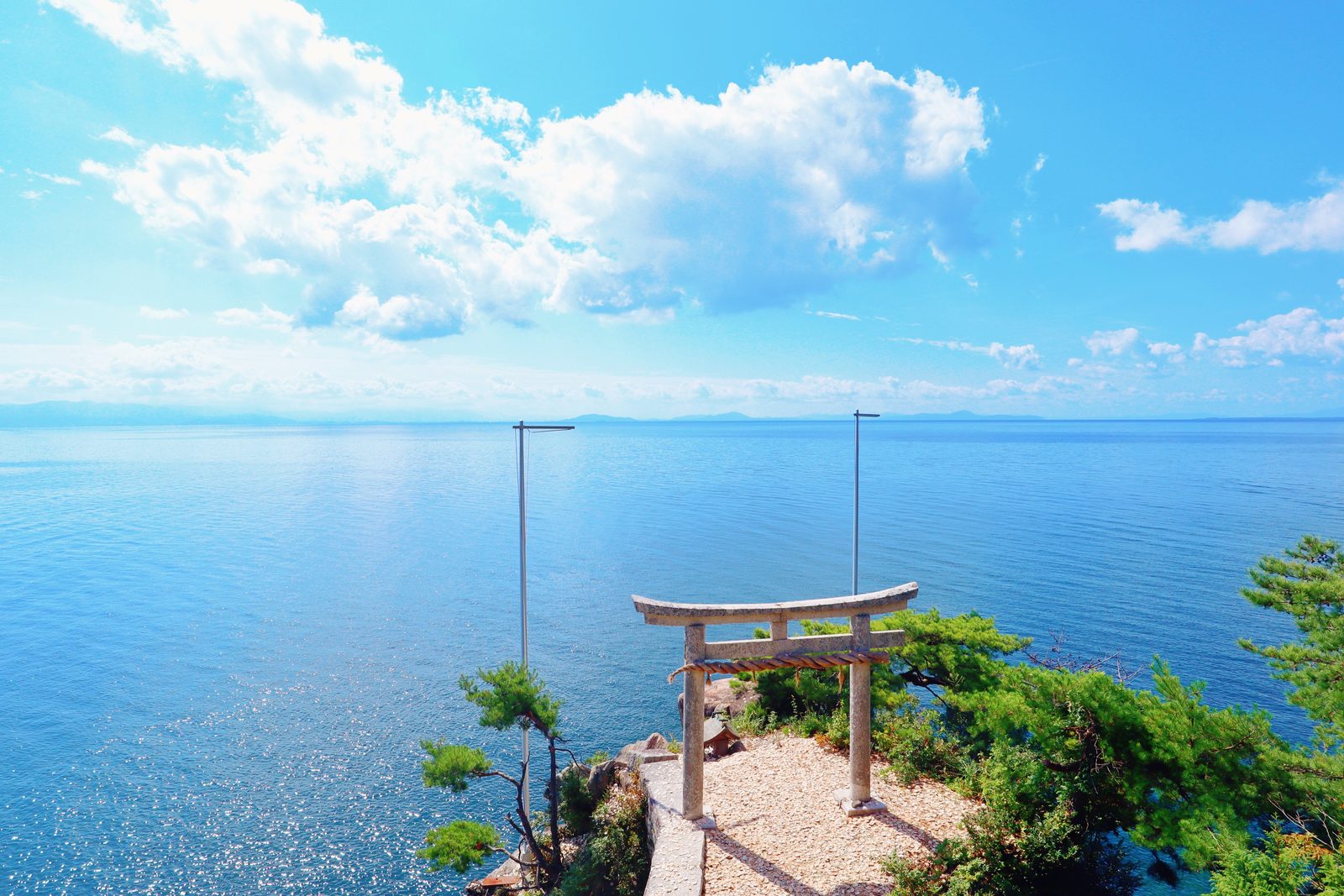 Stone torii gate on a lakeside cliff edge overlooking the wide, calm expanse of Lake Biwa, framed by pine branches