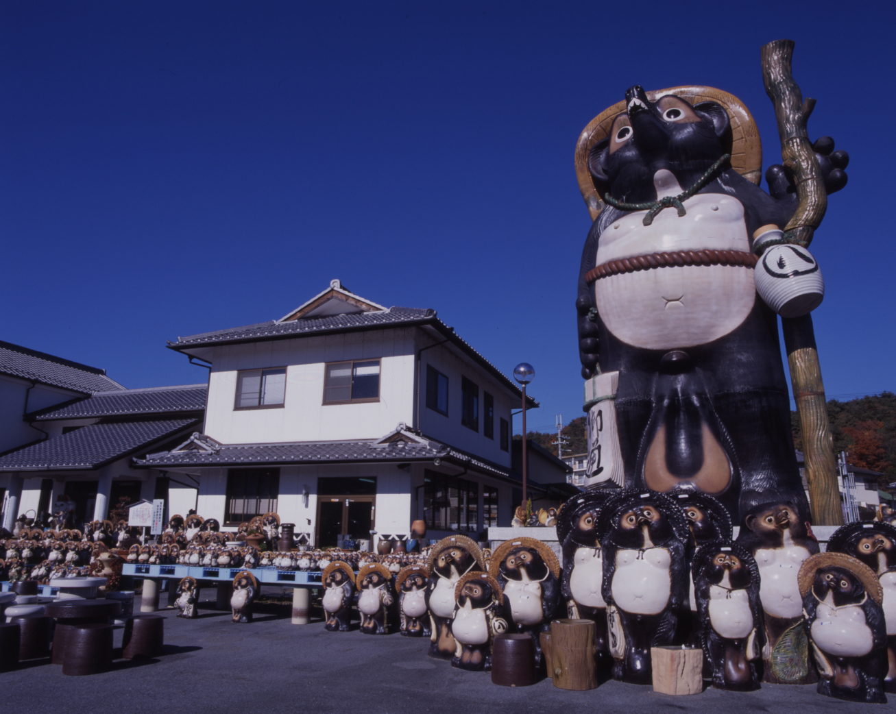 A large ceramic tanuki figure holding a staff beside a crowd of smaller tanuki figures lined up outside a white two-storey pottery shop.