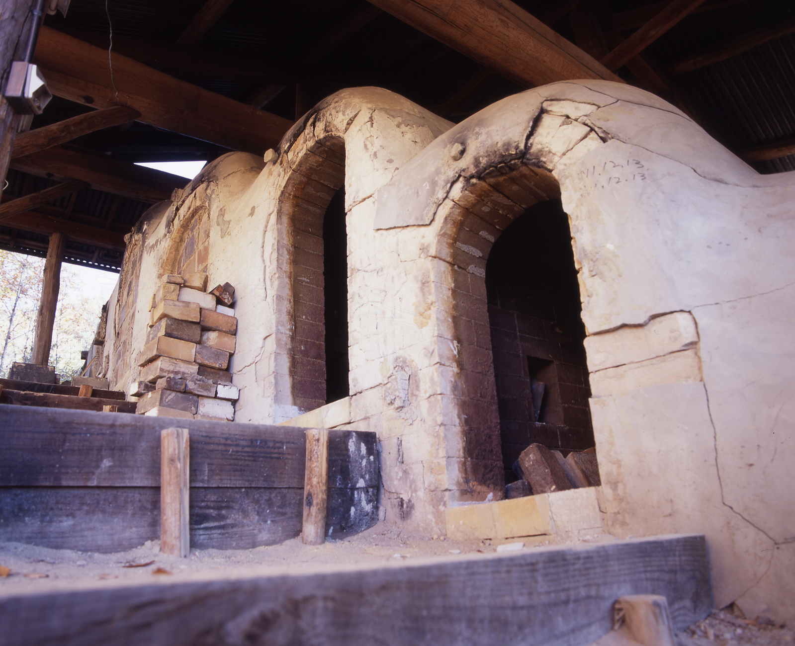 Two arched fire-mouth openings of a clay-and-brick climbing kiln chamber under a timber roof, with stacked kiln bricks in front.