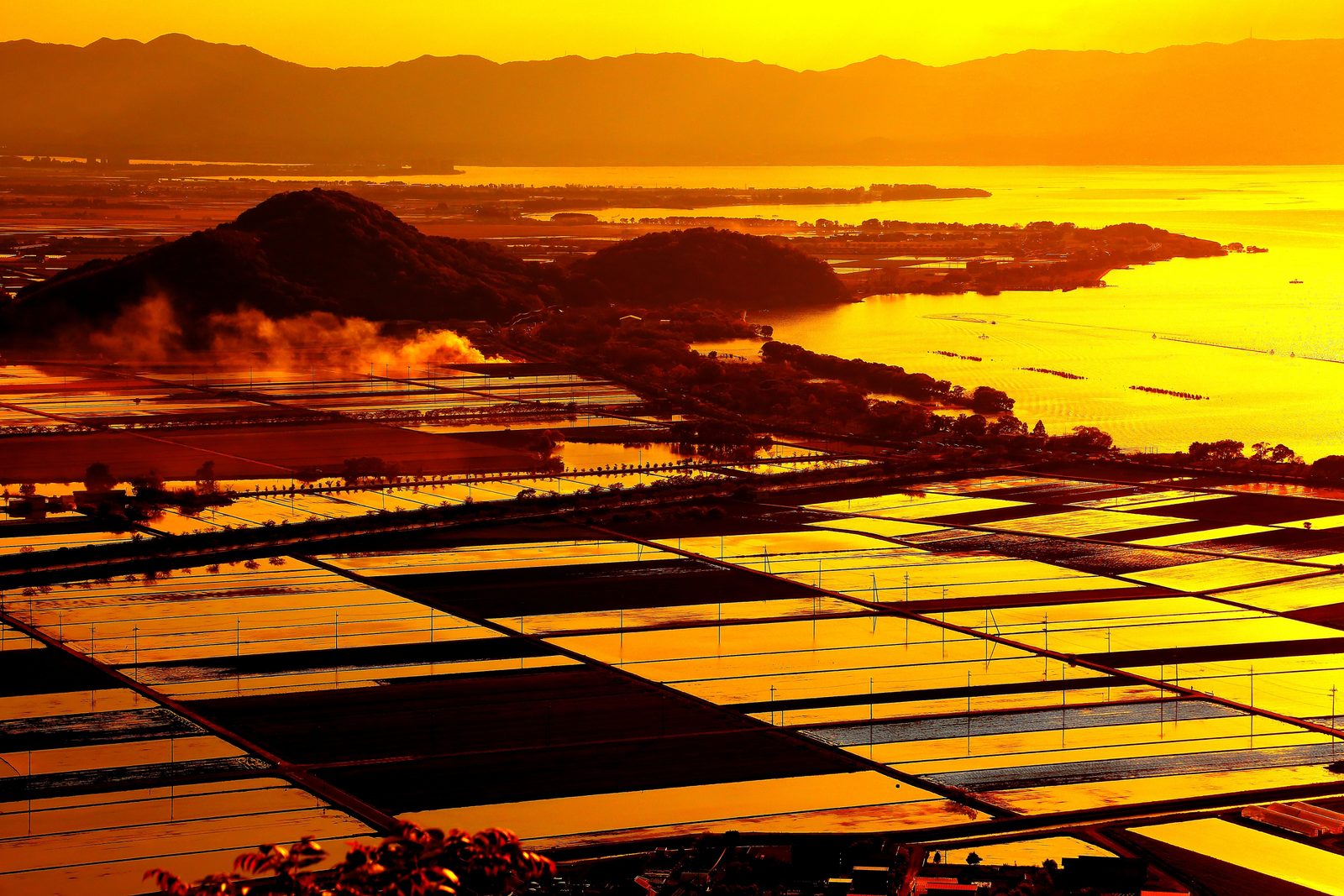 Flooded rice paddies on the lakeside plain of Omihachiman glowing orange at sunset, with a wooded hill, the lake and distant mountains beyond