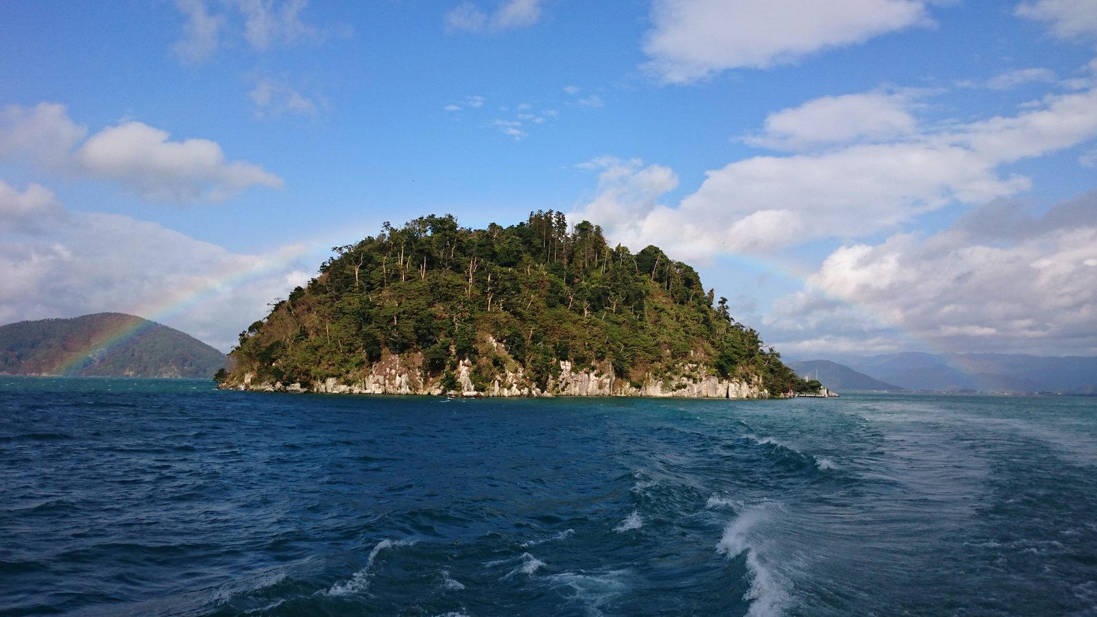 Chikubushima seen across the wake of a boat, a forested island with pale rock cliffs and a rainbow arching behind it