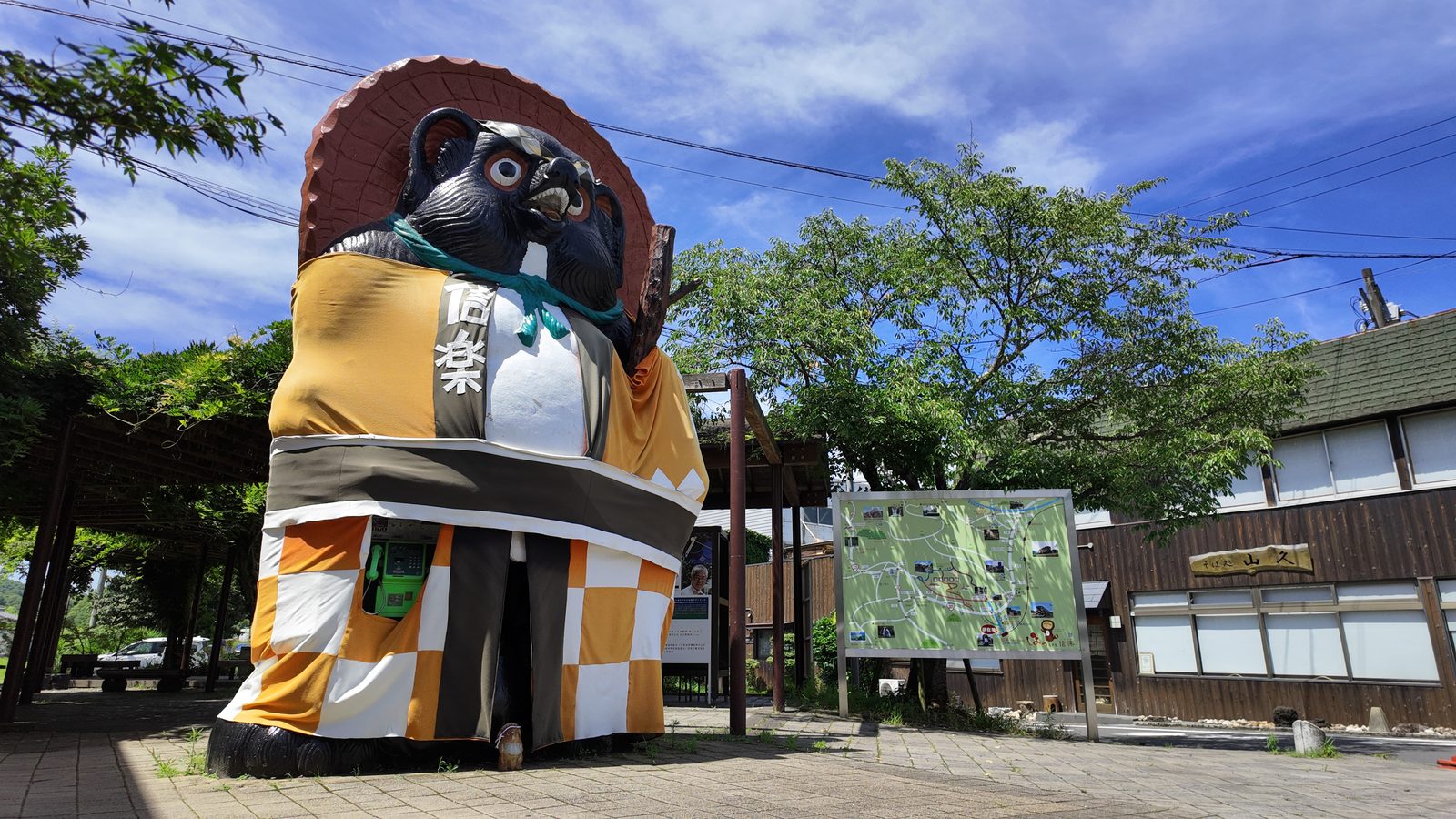 A giant ceramic tanuki figure wearing a straw hat and an orange and white checked wrap, standing in a paved square under a blue sky with wooden buildings behind it.