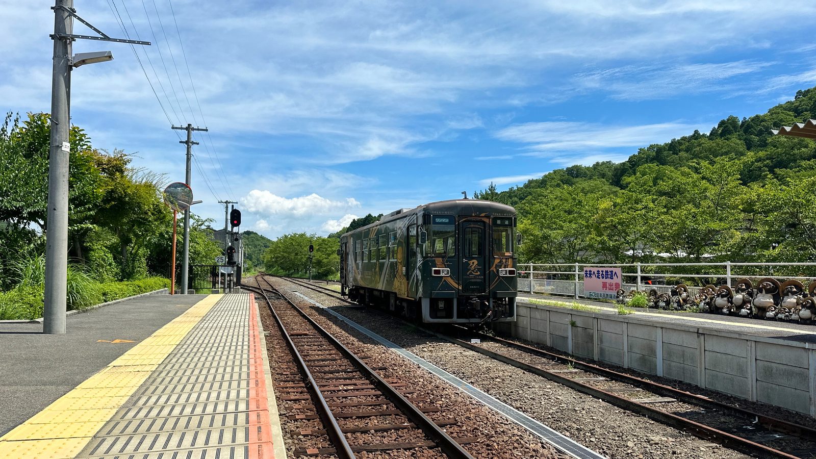 A dark green single-car train approaching a small station platform, with a row of tanuki figures lined up along the platform wall and wooded hills behind.