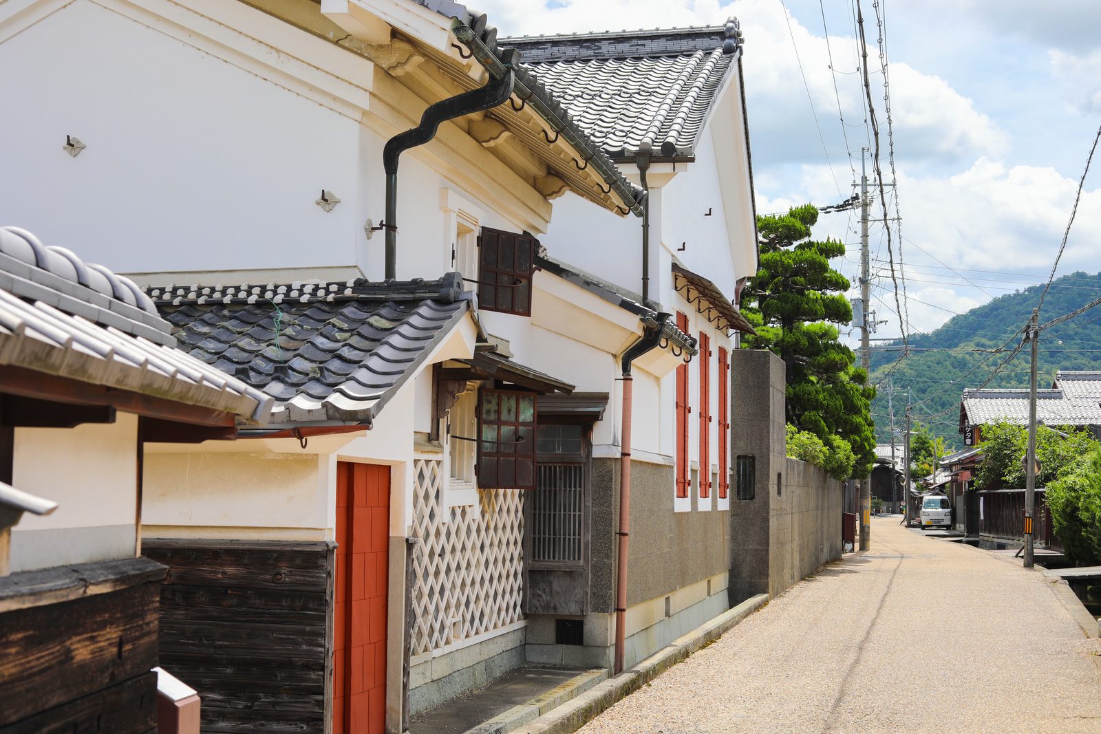 A quiet gravel street lined with white-plastered storehouses, one with a red door and a white diamond-lattice lower wall, with a pine tree and green hills in the distance.