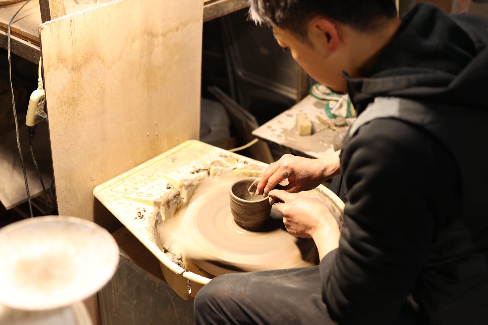A potter in a dark hoodie shaping a small clay bowl on a potter's wheel inside a workshop, seen from the side.