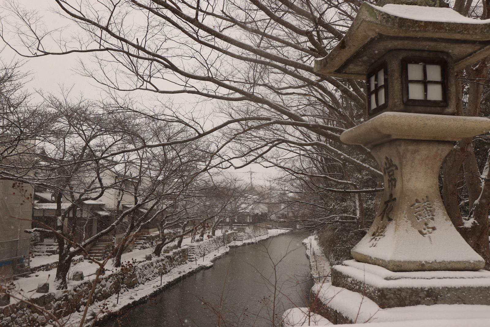 Snow-topped stone lantern beside a bridge looking down over the Hachimanbori canal, with bare trees and snow-covered stone walls along the dark water