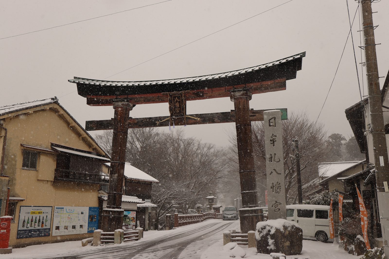 A large wooden torii and the stone marker of Himure Hachimangu Shrine on a snowy approach lined with old houses