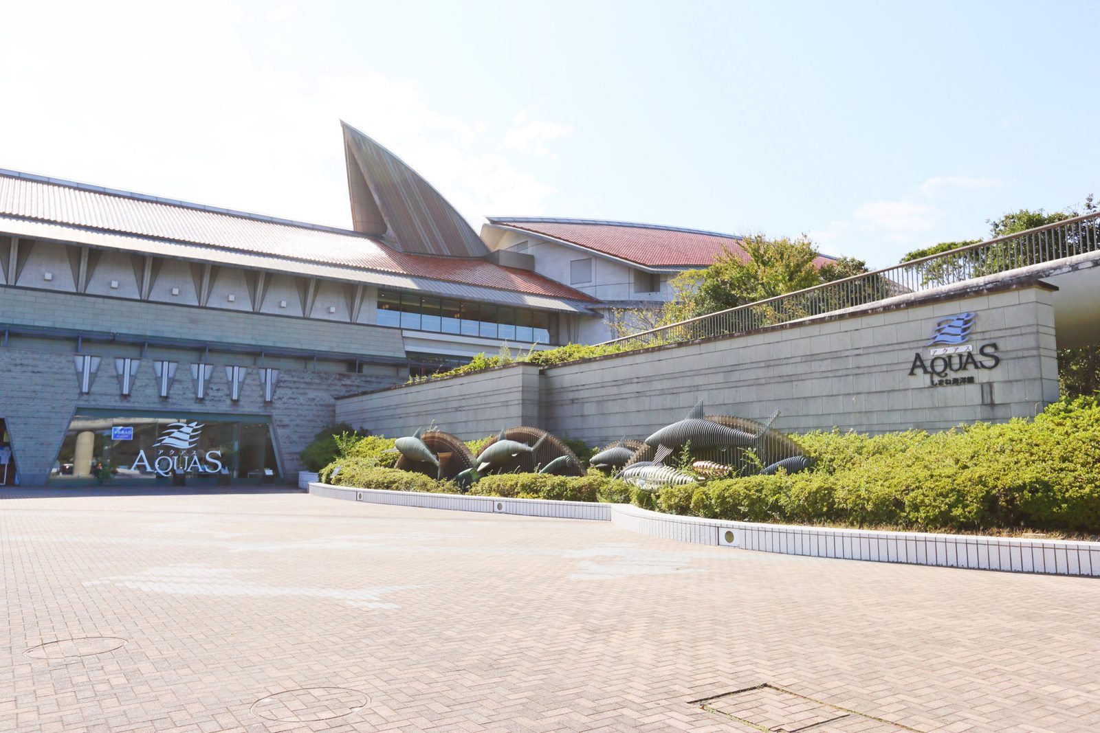 Entrance front of the Aquas aquarium building, with a paved plaza and fish-shaped metal sculptures along the wall