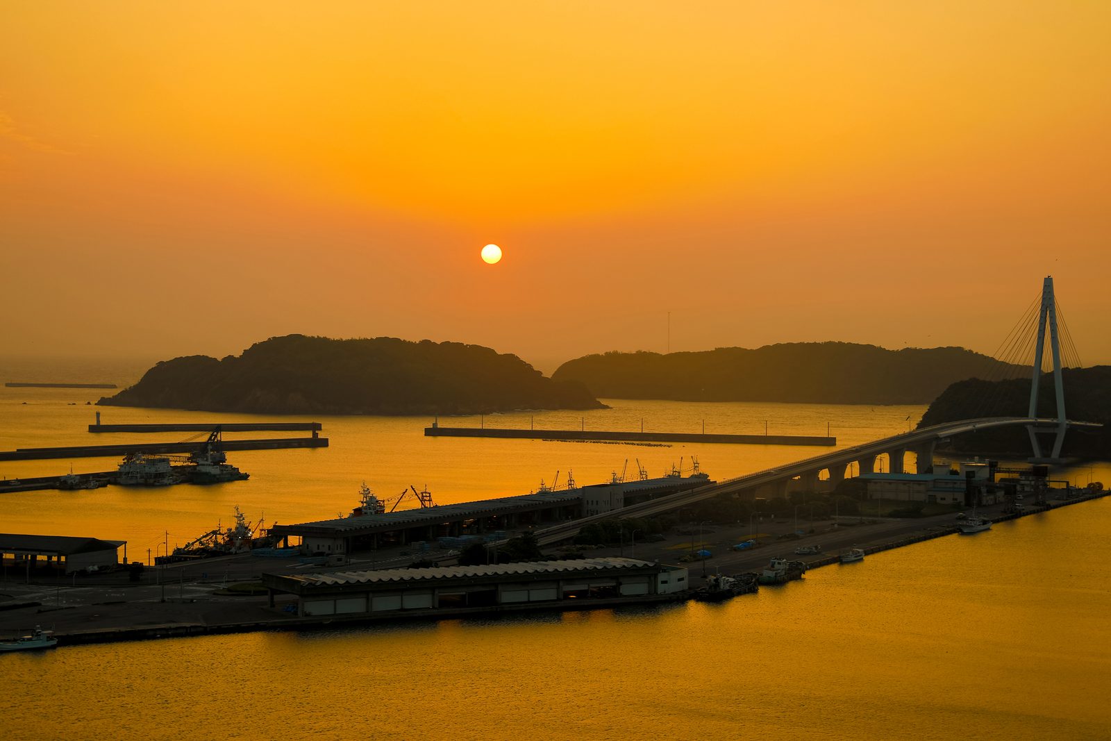 Sun low over a harbor at dusk, with wooded islands, breakwaters, fishing-port buildings and a cable-stayed bridge in orange light