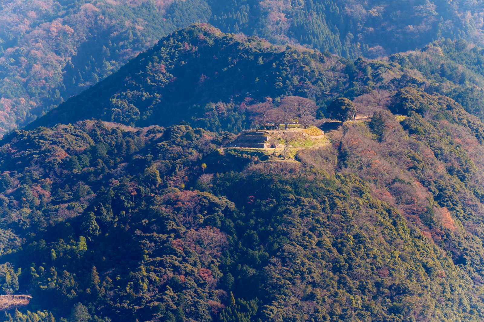 Wooded mountain ridge with the stone terraces of a castle site on its crest, seen from across the valley