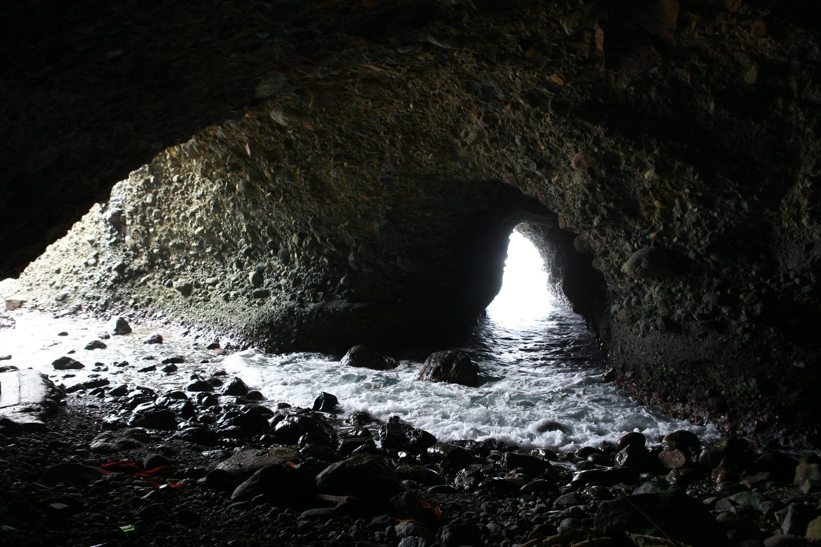 Looking out from inside a dark sea cave, where waves wash over dark stones toward a bright arched opening