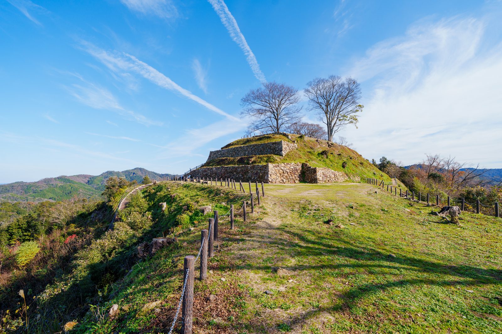 Stone-walled terraces of a hilltop castle site with two bare trees against a blue sky